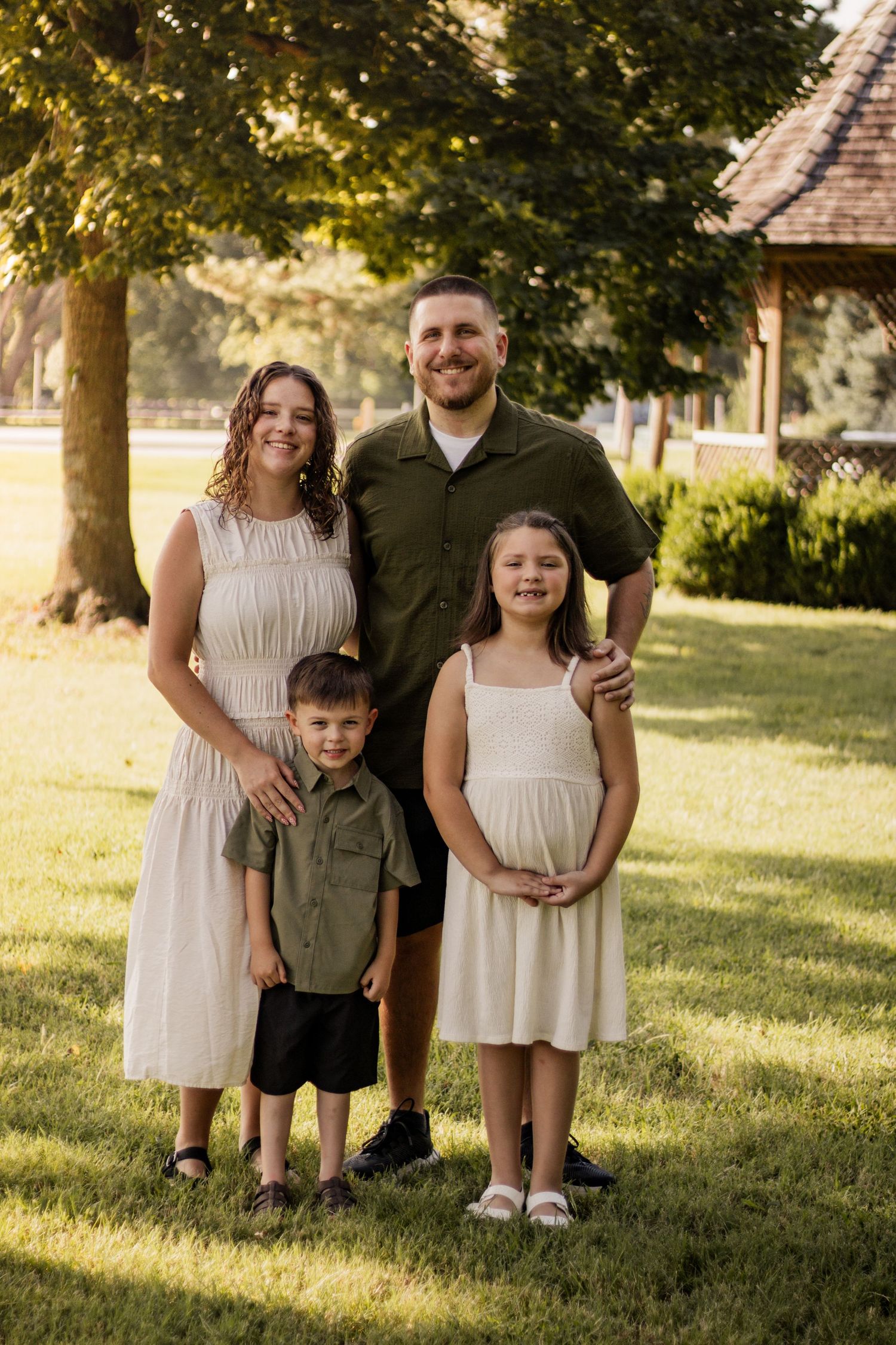 A smiling family of four poses together on a green lawn with trees and a building in the background.