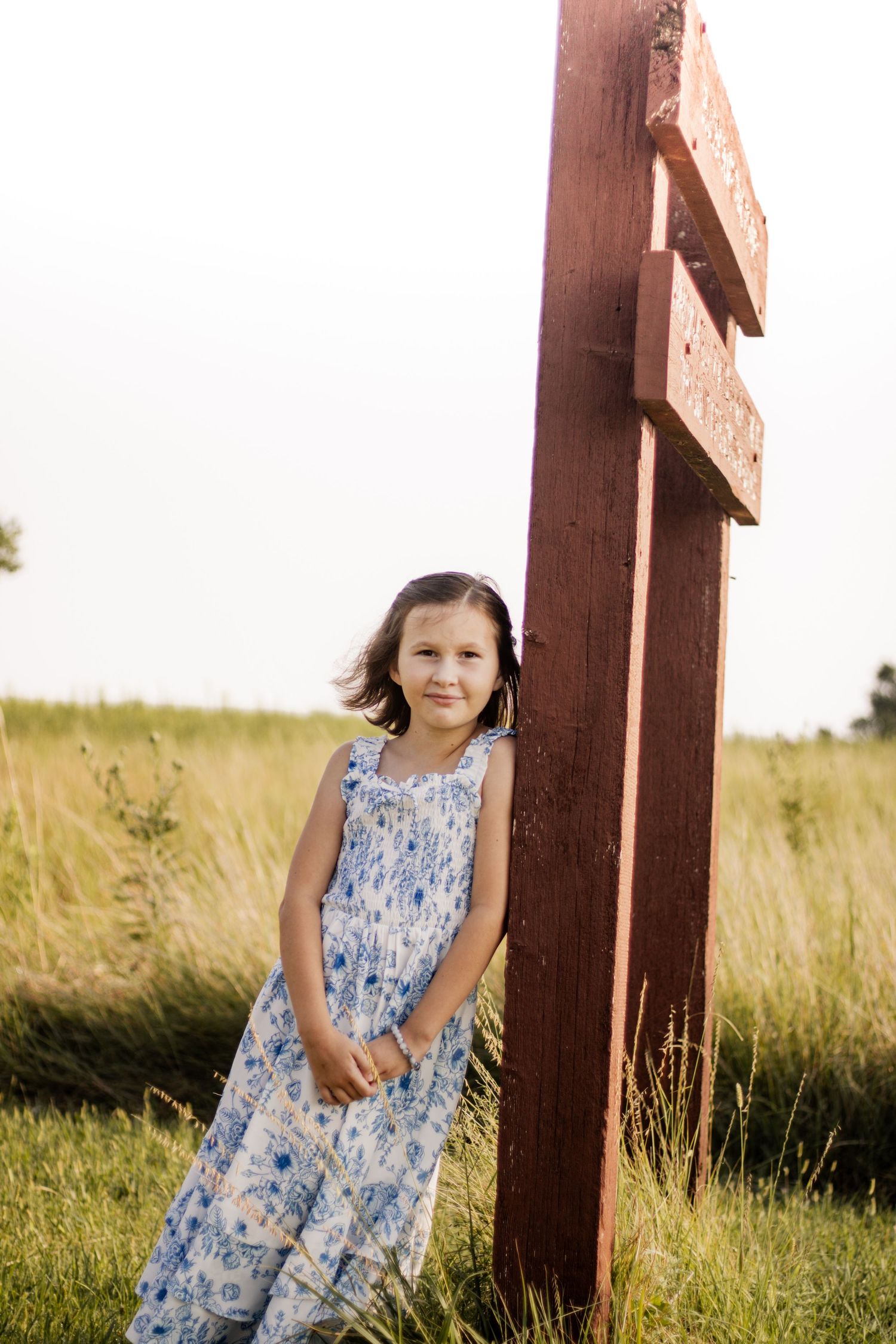 Young girl in floral dress standing beside a rustic red wooden post in a sunny green meadow.