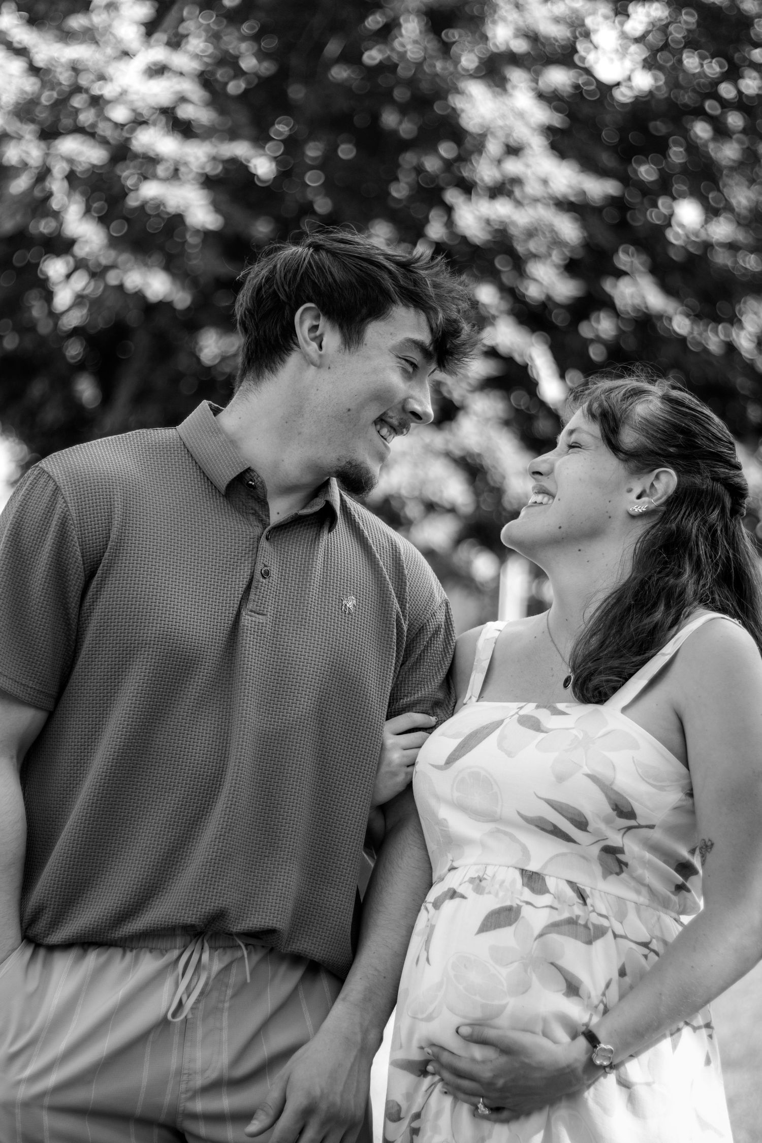 Black and white photo of a smiling pregnant couple gazing at each other outdoors with trees in the background.