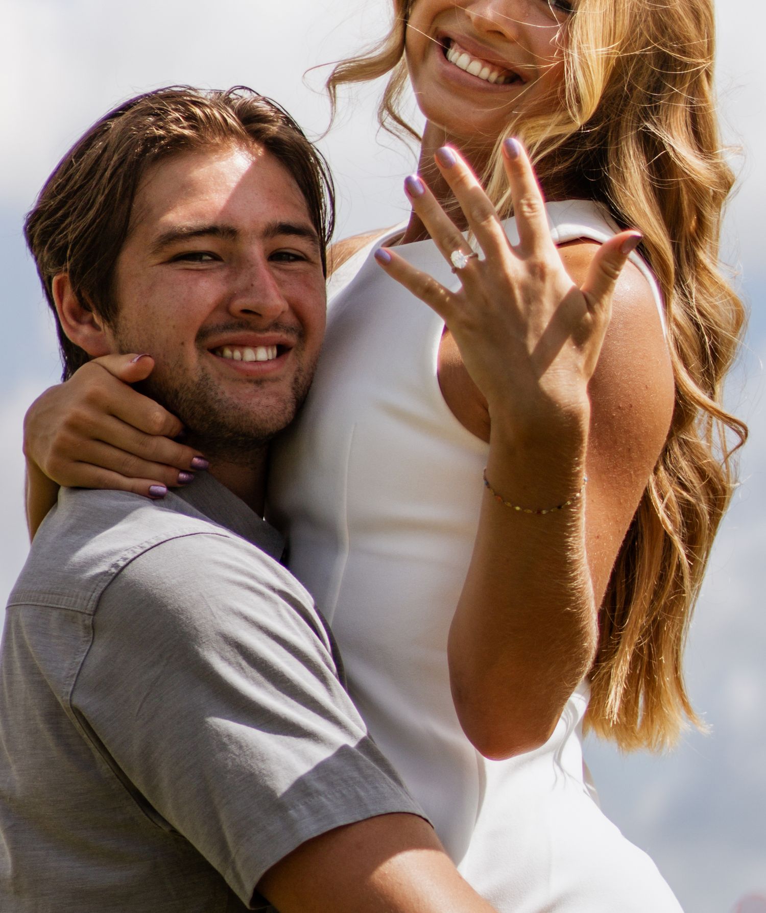 Happy couple celebrating engagement outdoors, woman showing diamond ring while smiling, man hugging her from behind.