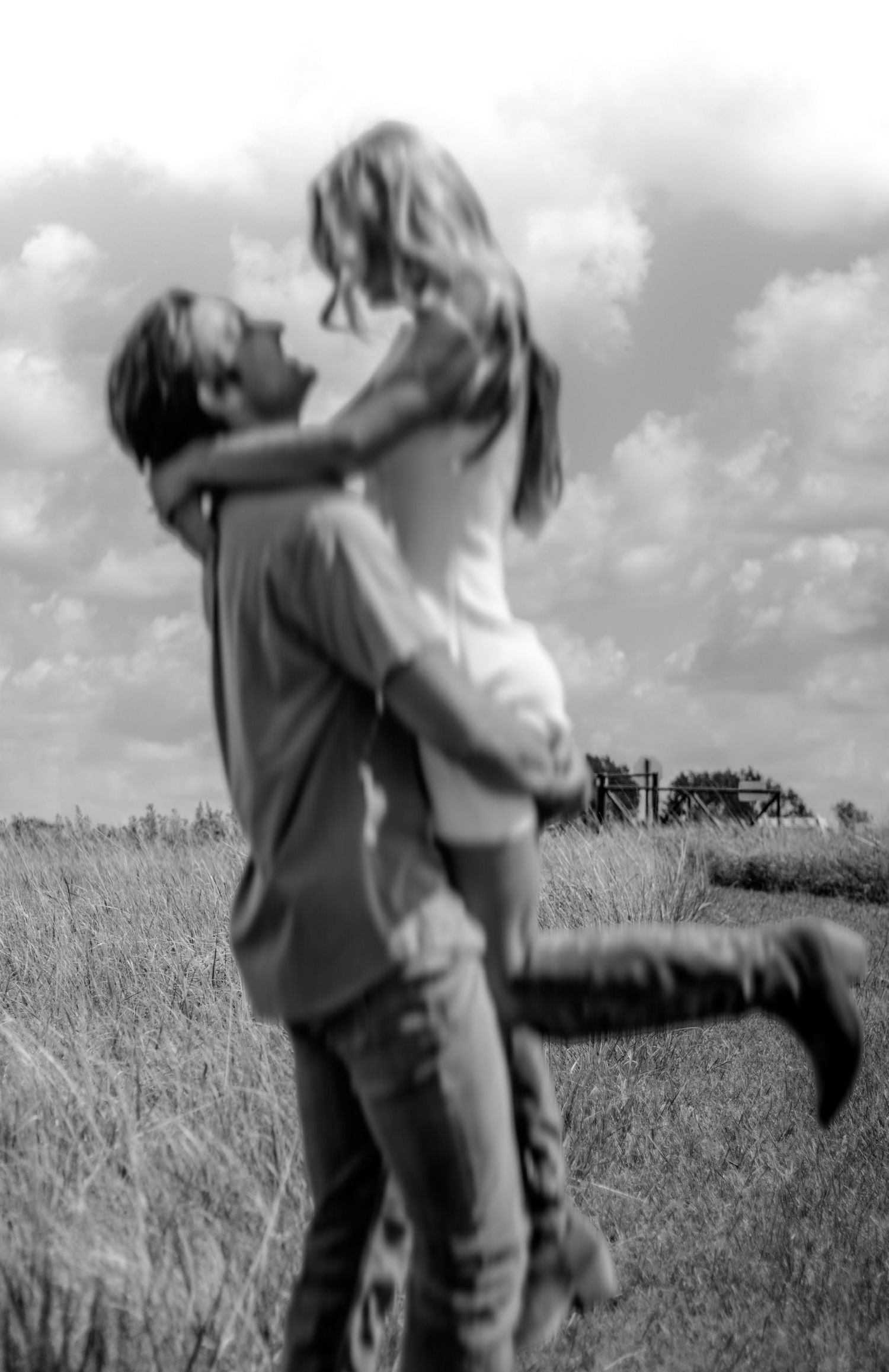 Black and white romantic photo of couple embracing in a field, woman lifted off ground kissing man.
