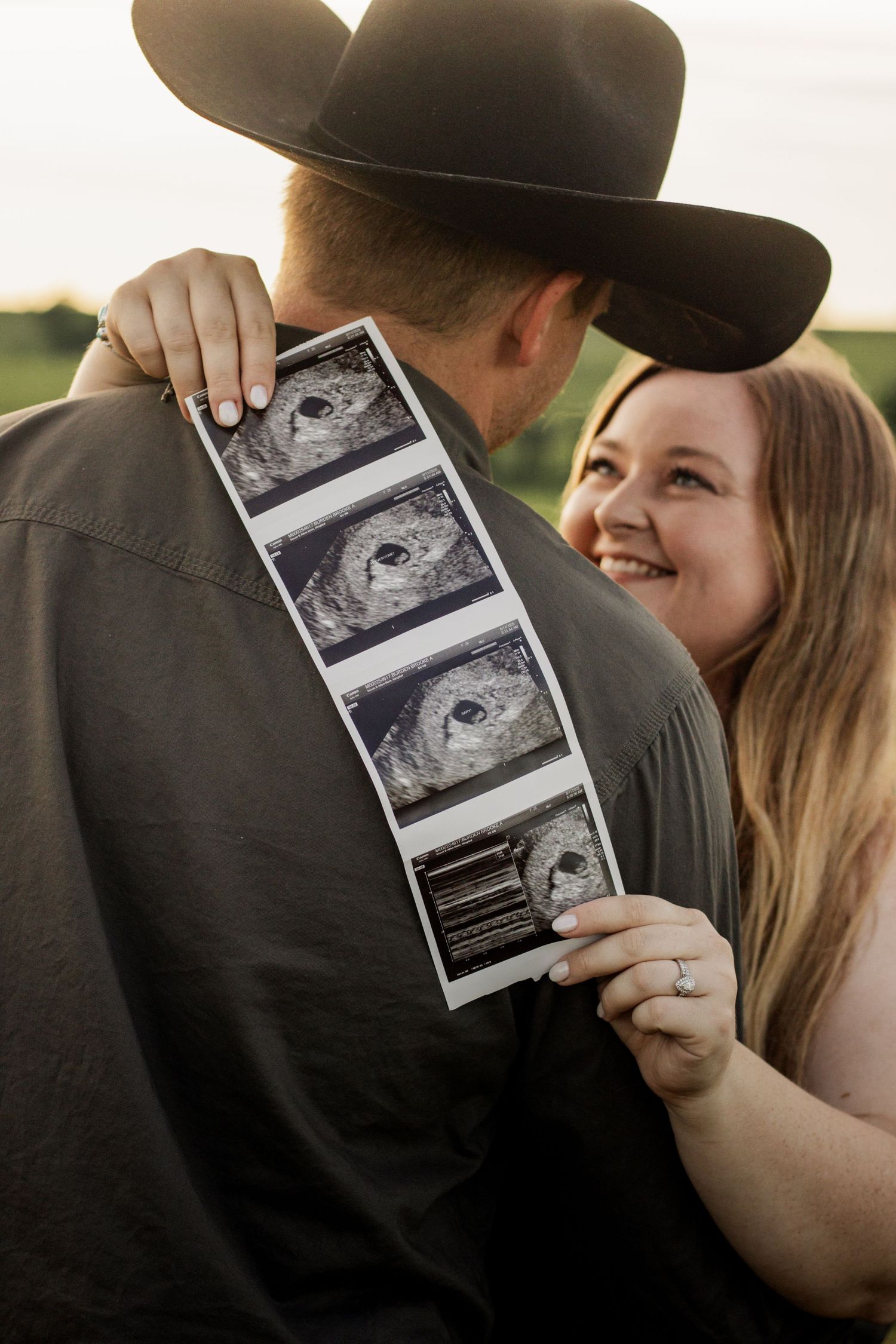 Smiling woman holds ultrasound photos behind man in cowboy hat during outdoor pregnancy announcement photoshoot.