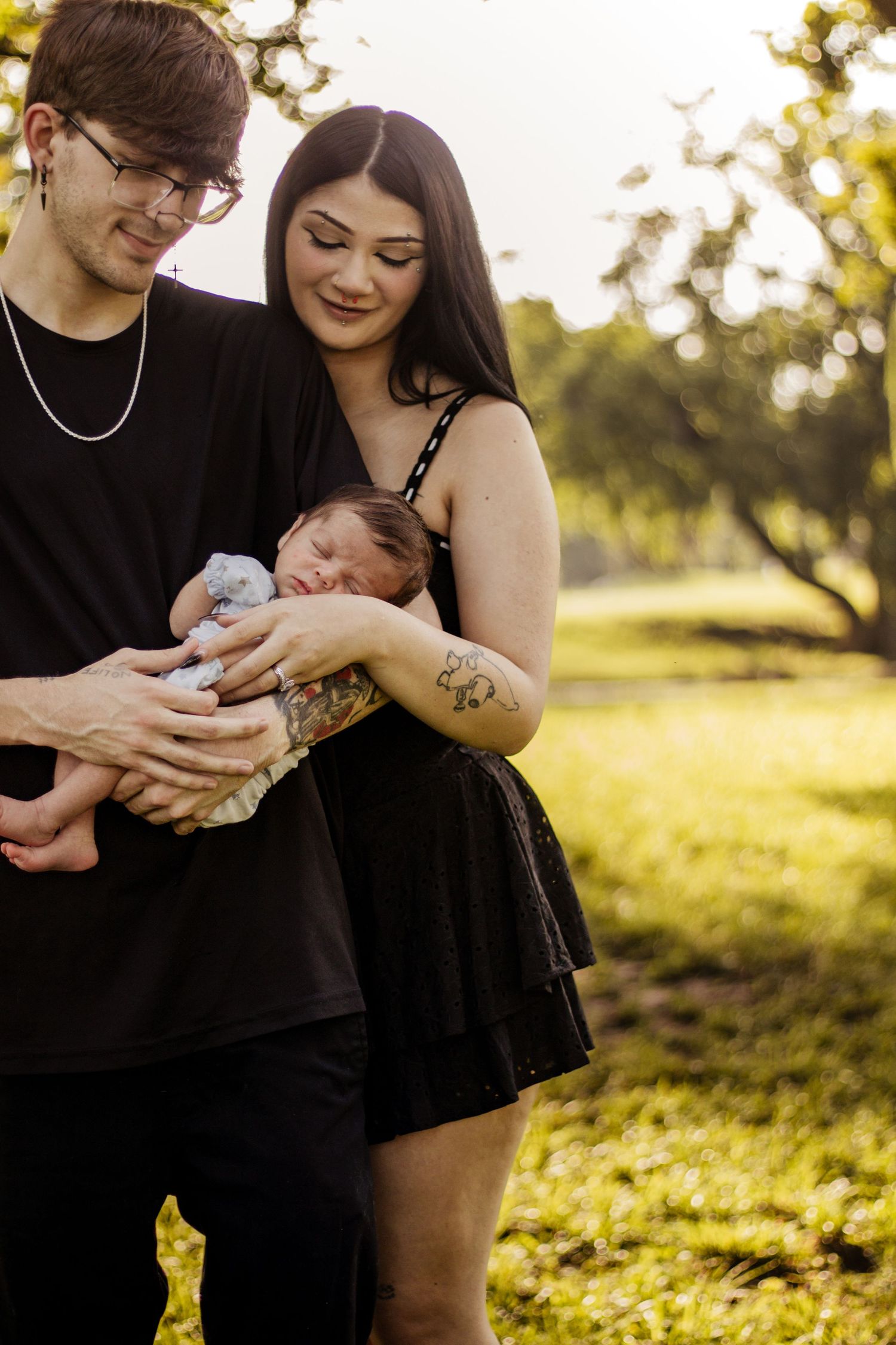 Young couple in black outfits cradling their newborn baby outdoors in a sunlit park setting.
