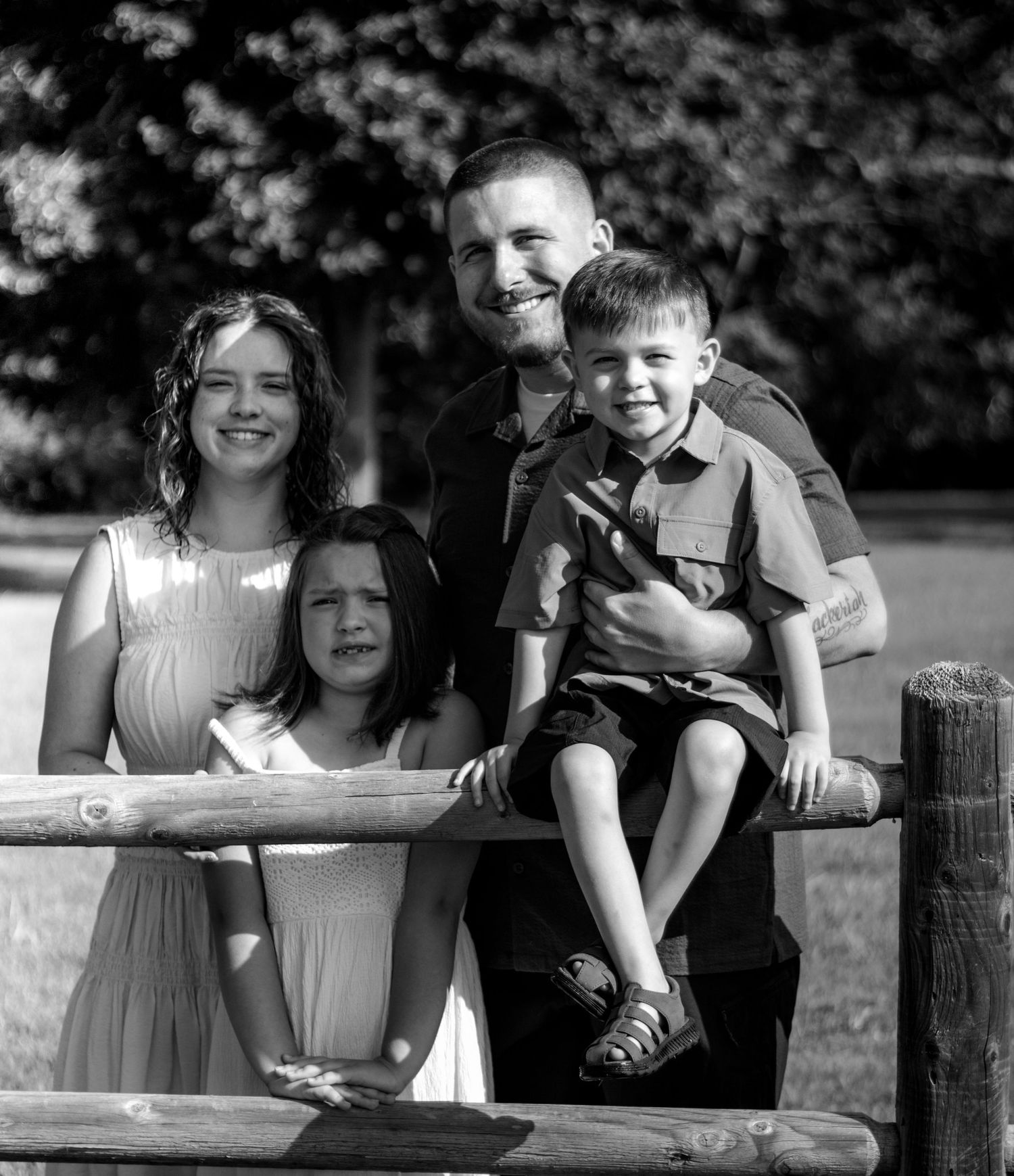 Black and white family portrait of parents with two young children posing by a rustic wooden fence outdoors.