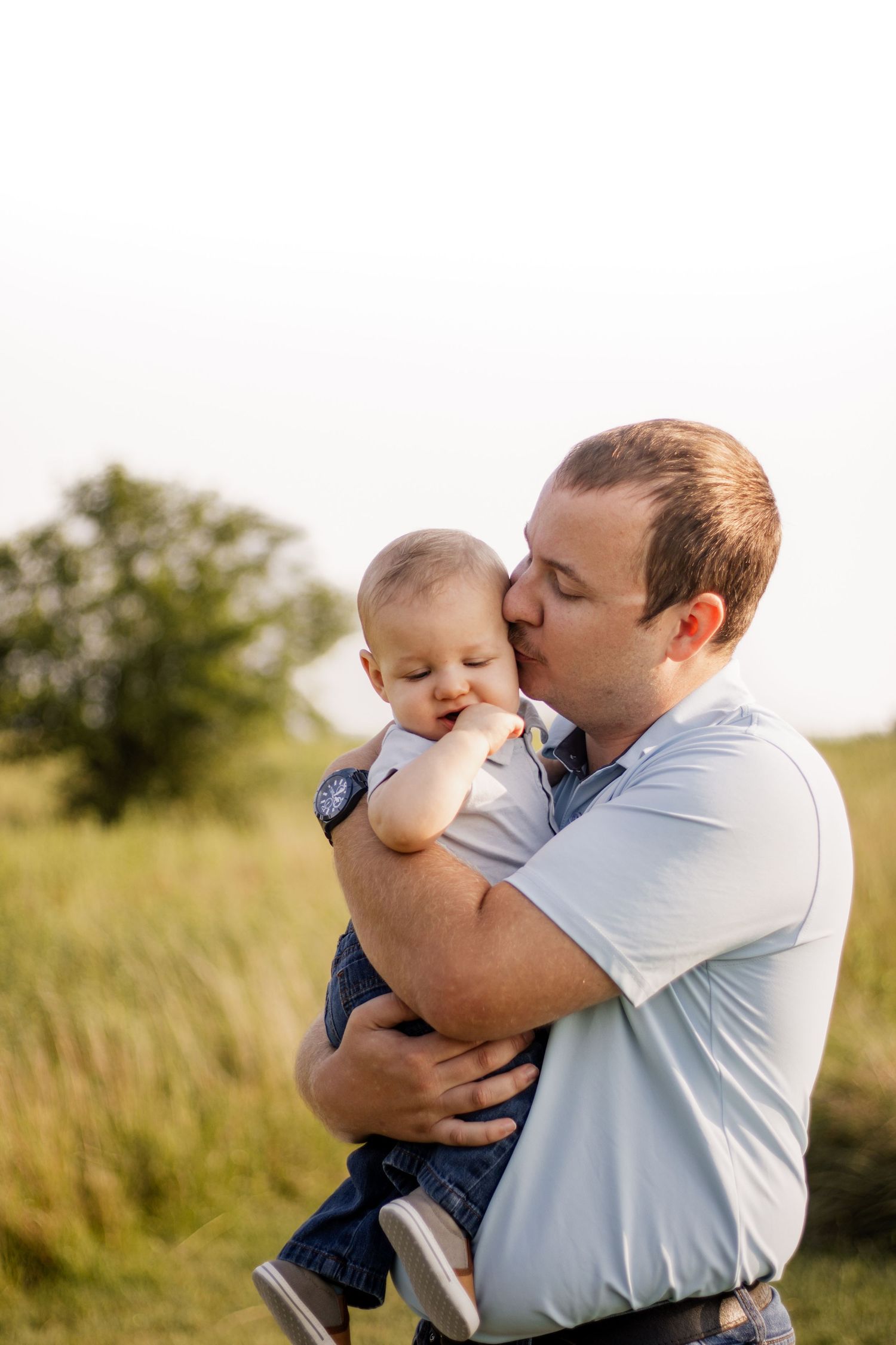 A father tenderly kisses his baby while holding them in a sunlit green field.