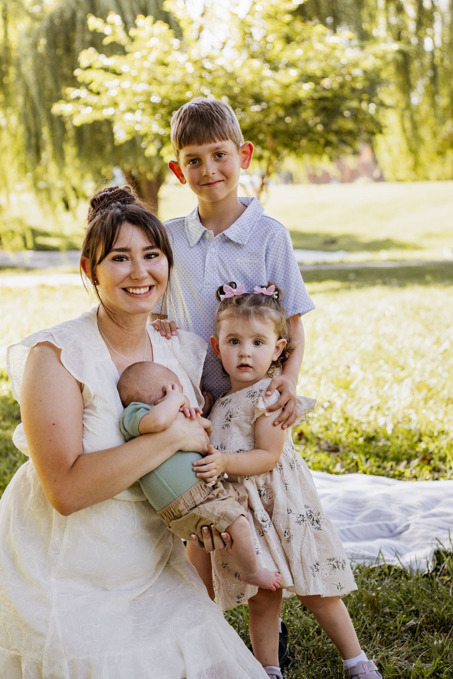 A smiling mother in a white dress holds a newborn while posing outdoors with her toddler daughter and young son.