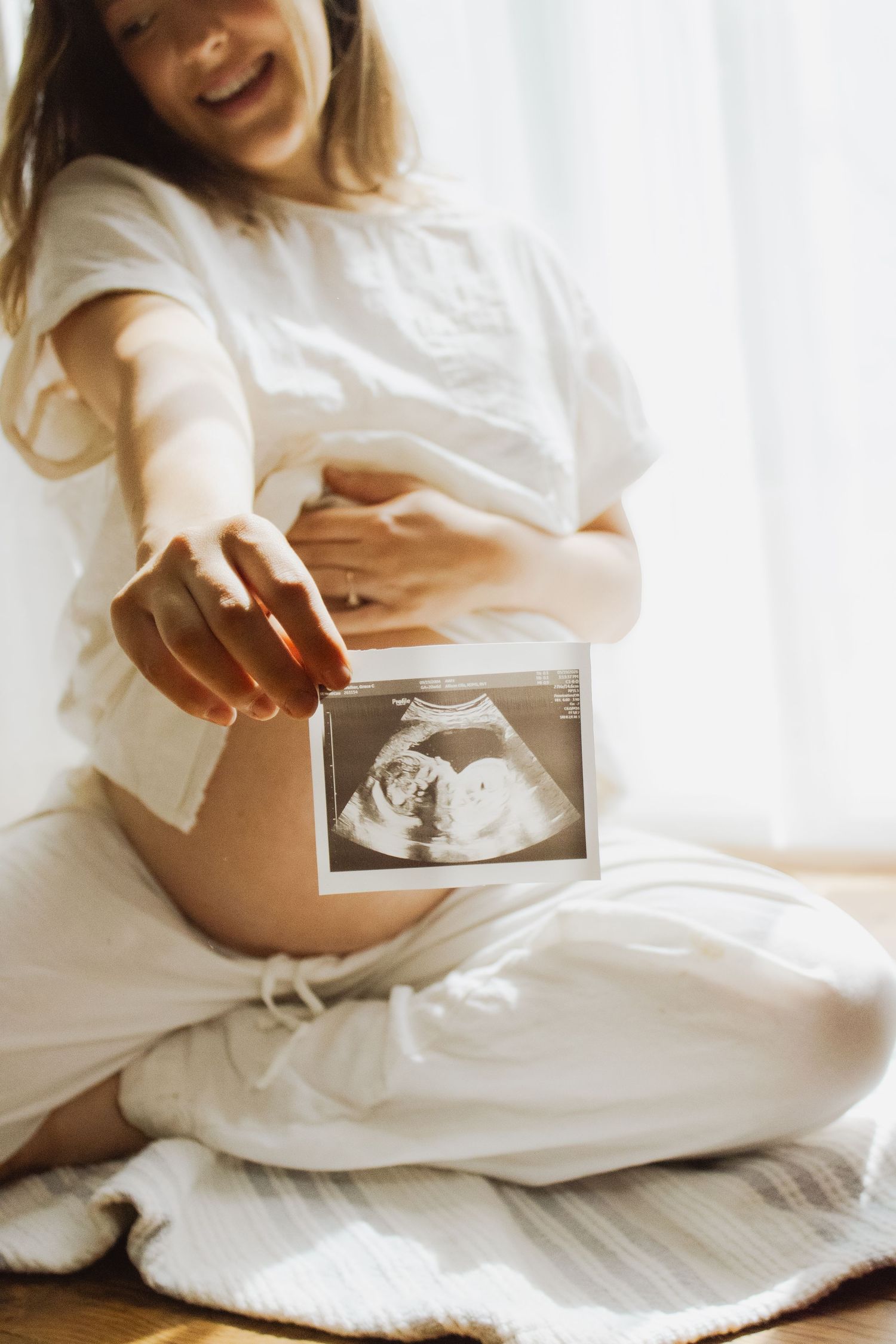 Laughing pregnant woman in white linen sitting cross-legged holding ultrasound scan near baby bump.