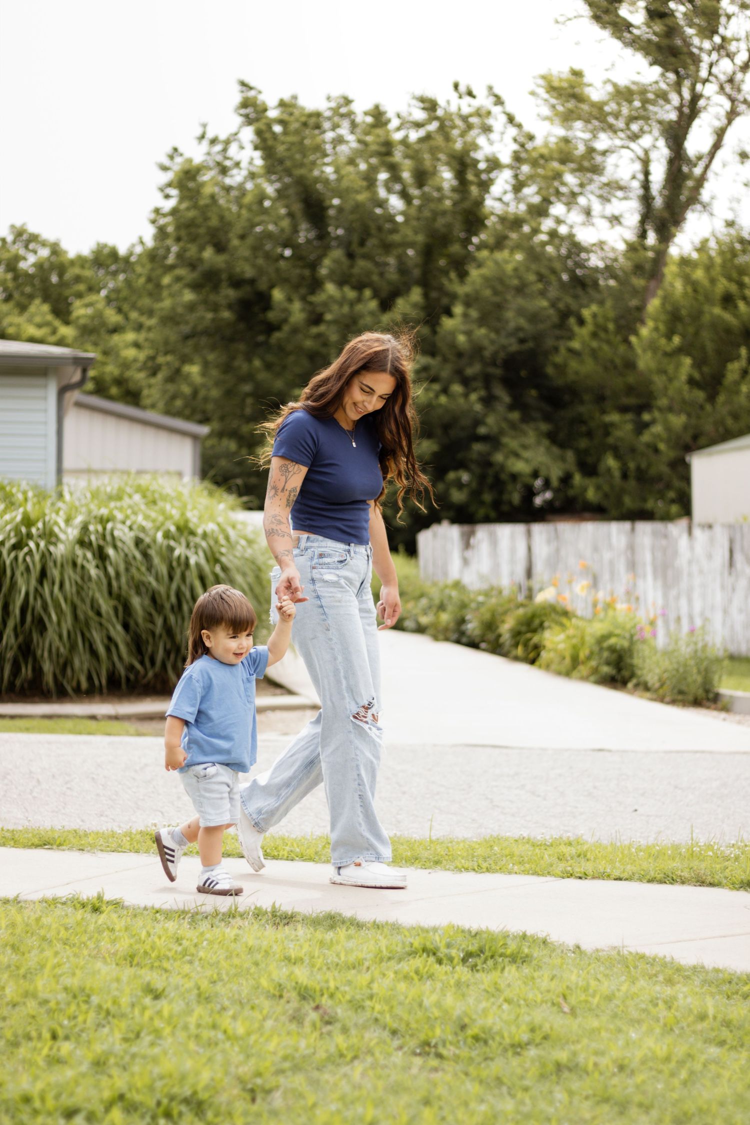 A young mother holds her toddler's hand while walking together on a sunny sidewalk in a residential neighborhood.