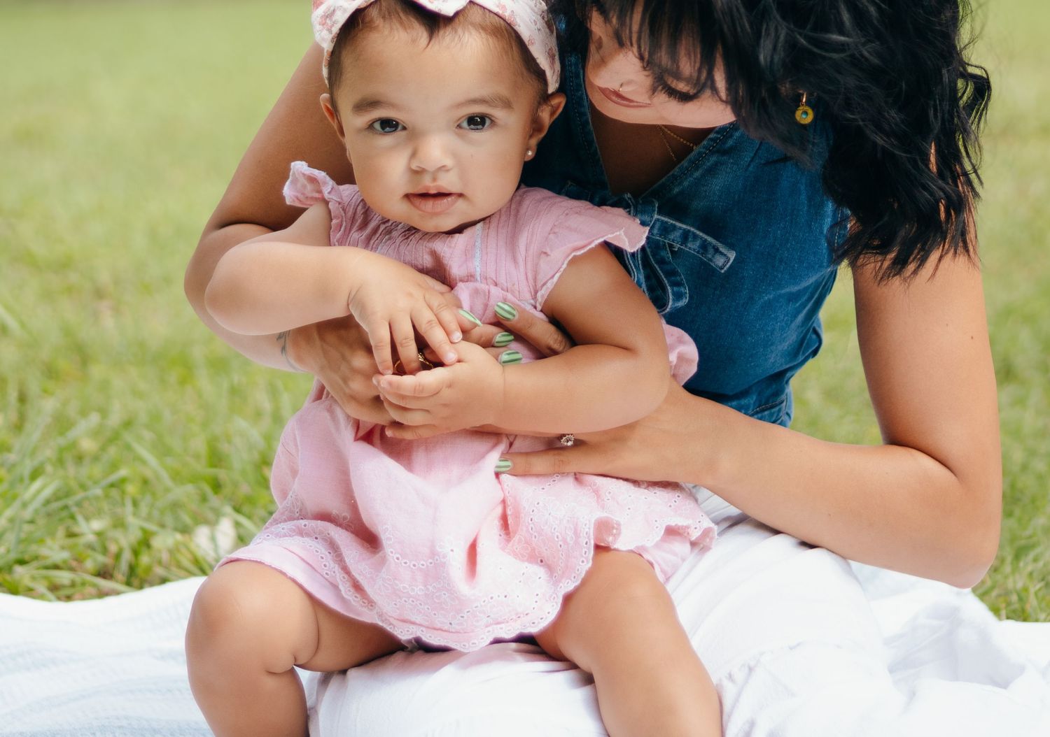 A mother in denim overalls holds her baby girl in a pink dress on a white blanket outdoors.