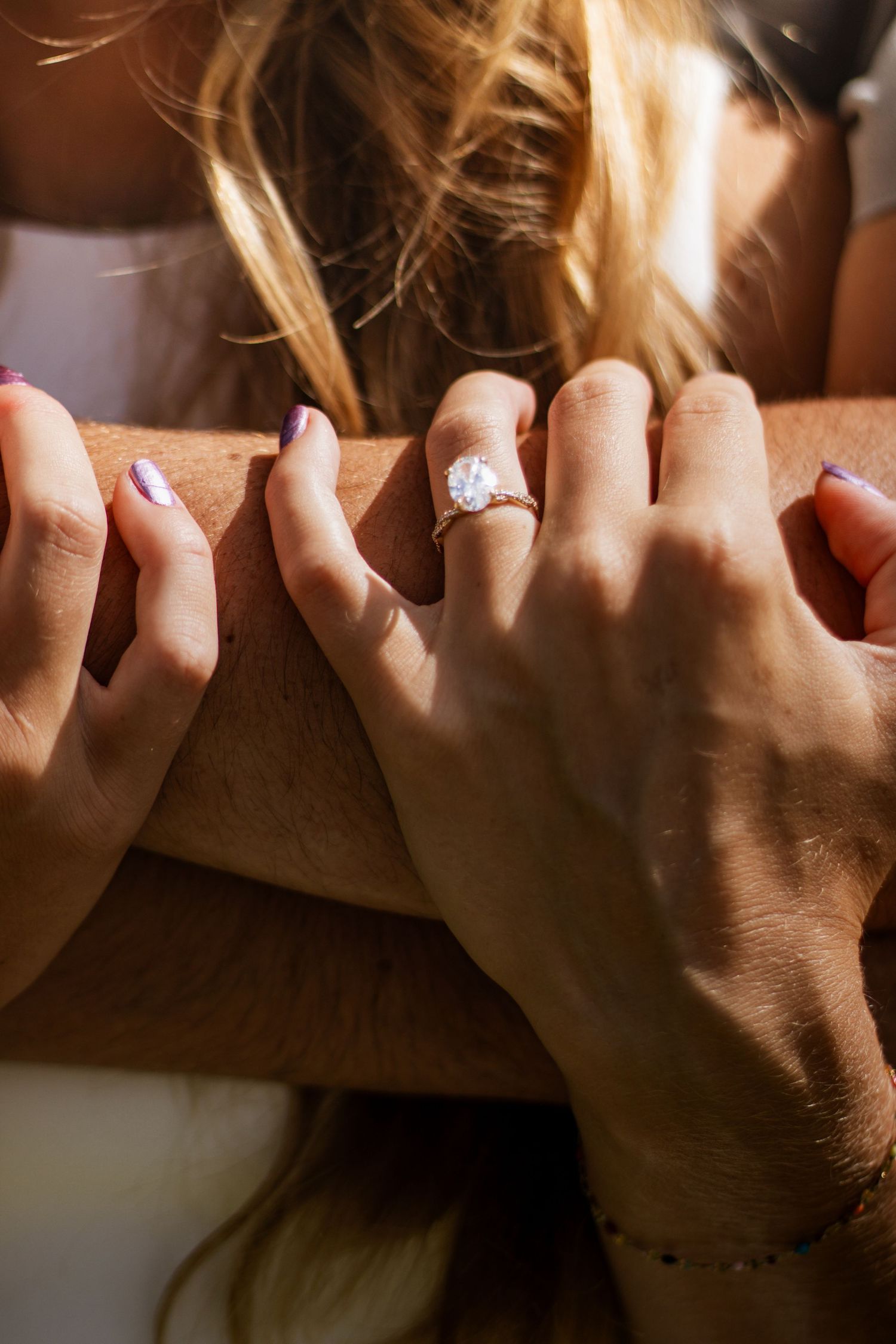 Close-up of hands resting on a wooden surface, featuring a stunning diamond engagement ring with purple manicured nails.