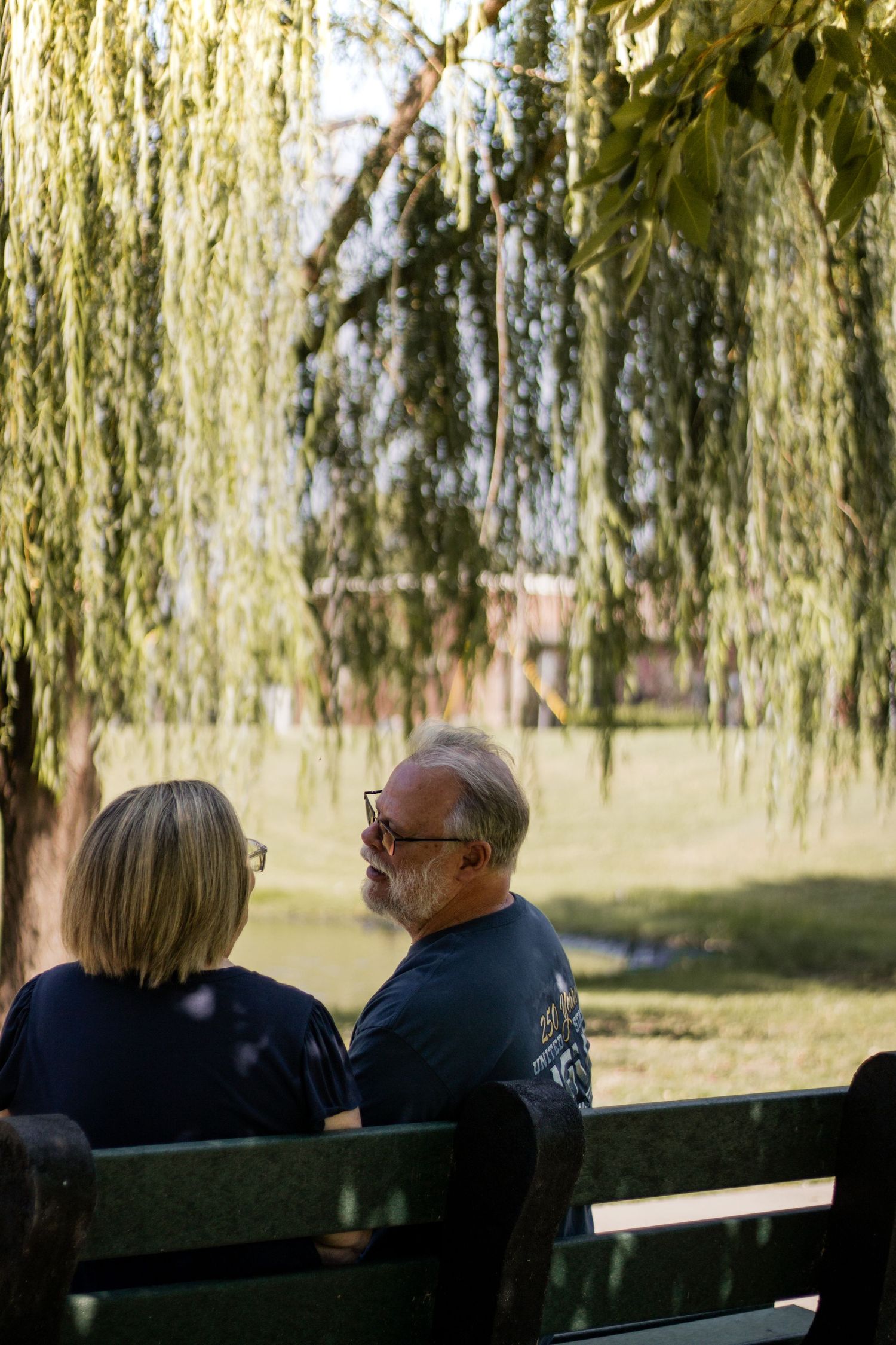 Older couple sitting on a park bench under a weeping willow tree, enjoying a sunny day outdoors.