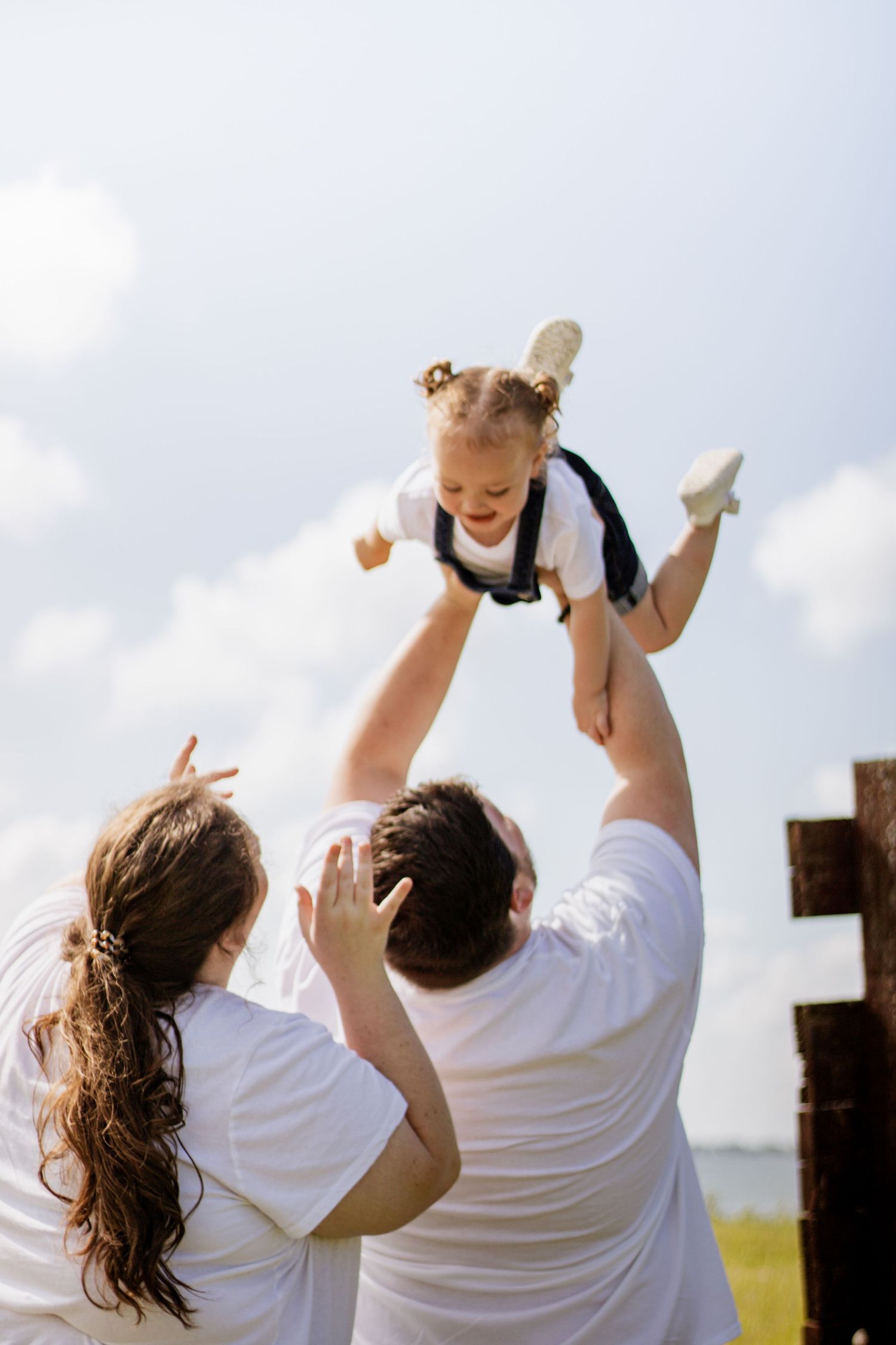 Parents joyfully lifting their toddler high in the air outdoors on a sunny day with blue sky background.