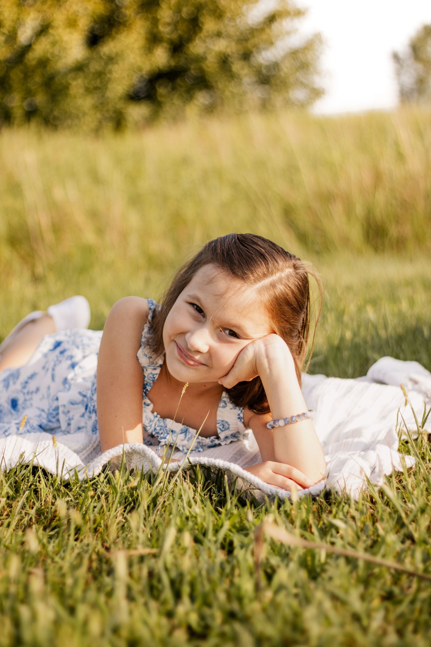 Young girl in floral dress lying on white blanket in sunny green field, smiling with hand on cheek.