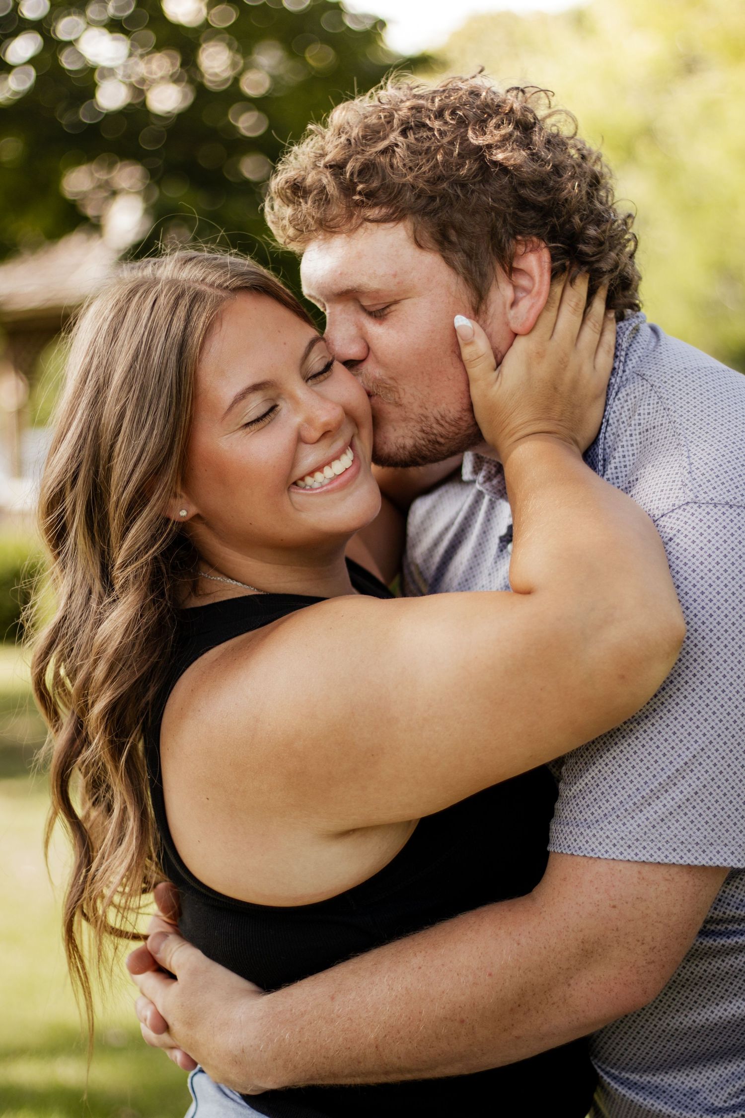 A happy couple sharing a tender kiss outdoors, with the woman smiling joyfully in a black top.