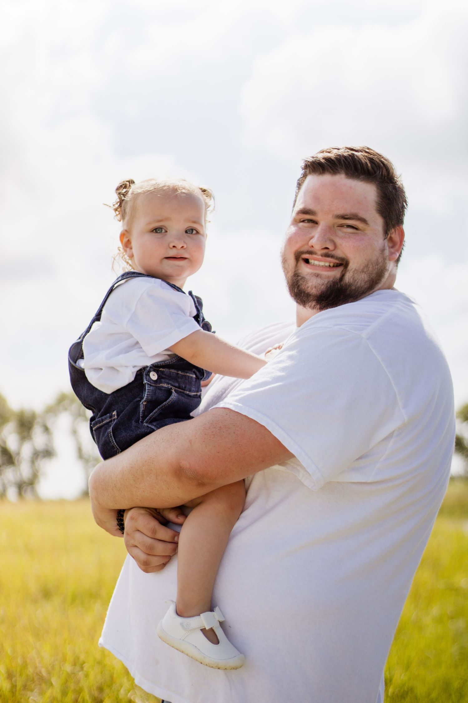 Smiling father holds toddler girl in denim overalls outdoors in a green field under a cloudy sky.