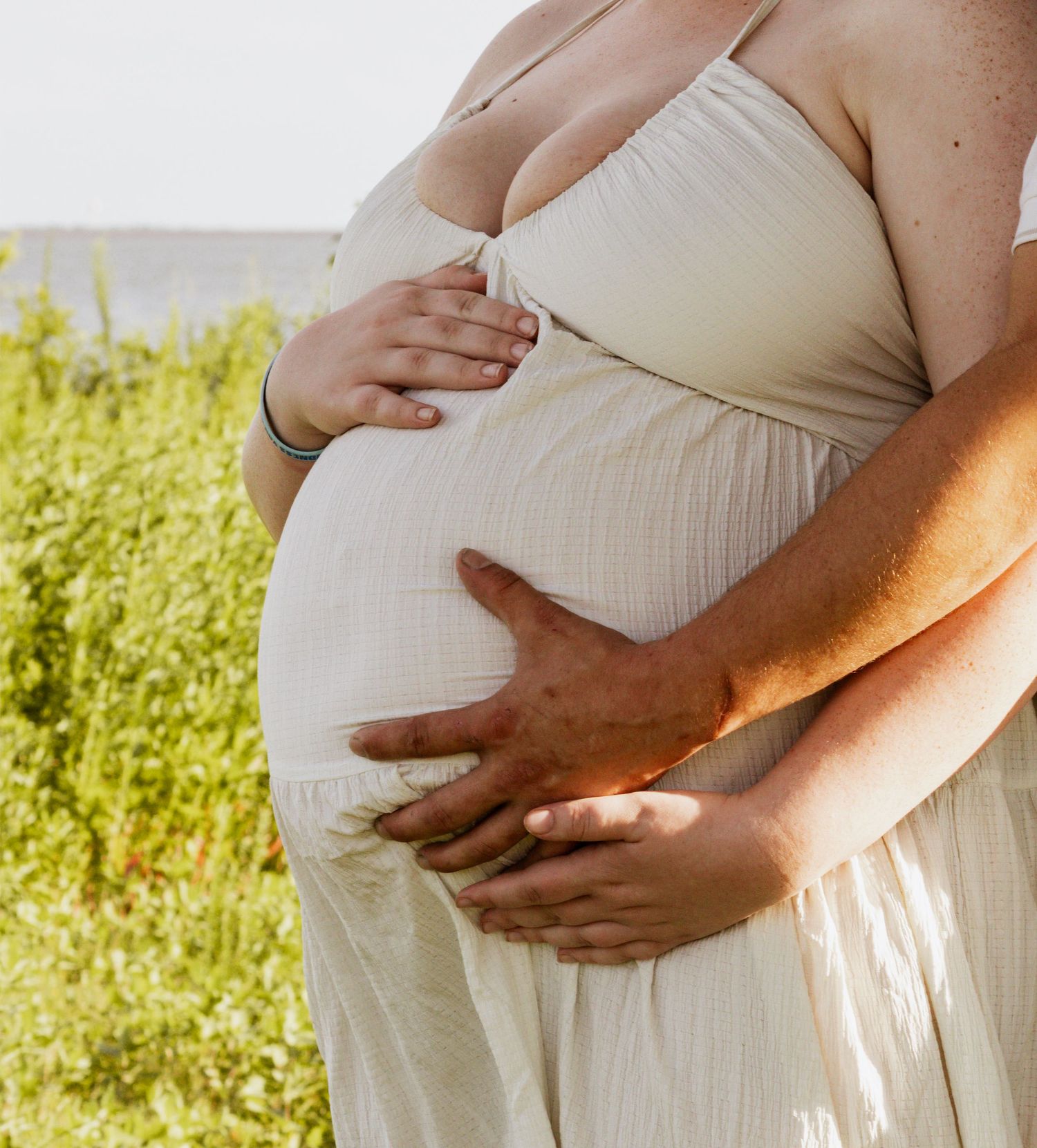 Pregnant woman in white dress cradling baby bump outdoors with partner's hands wrapped around her belly.
