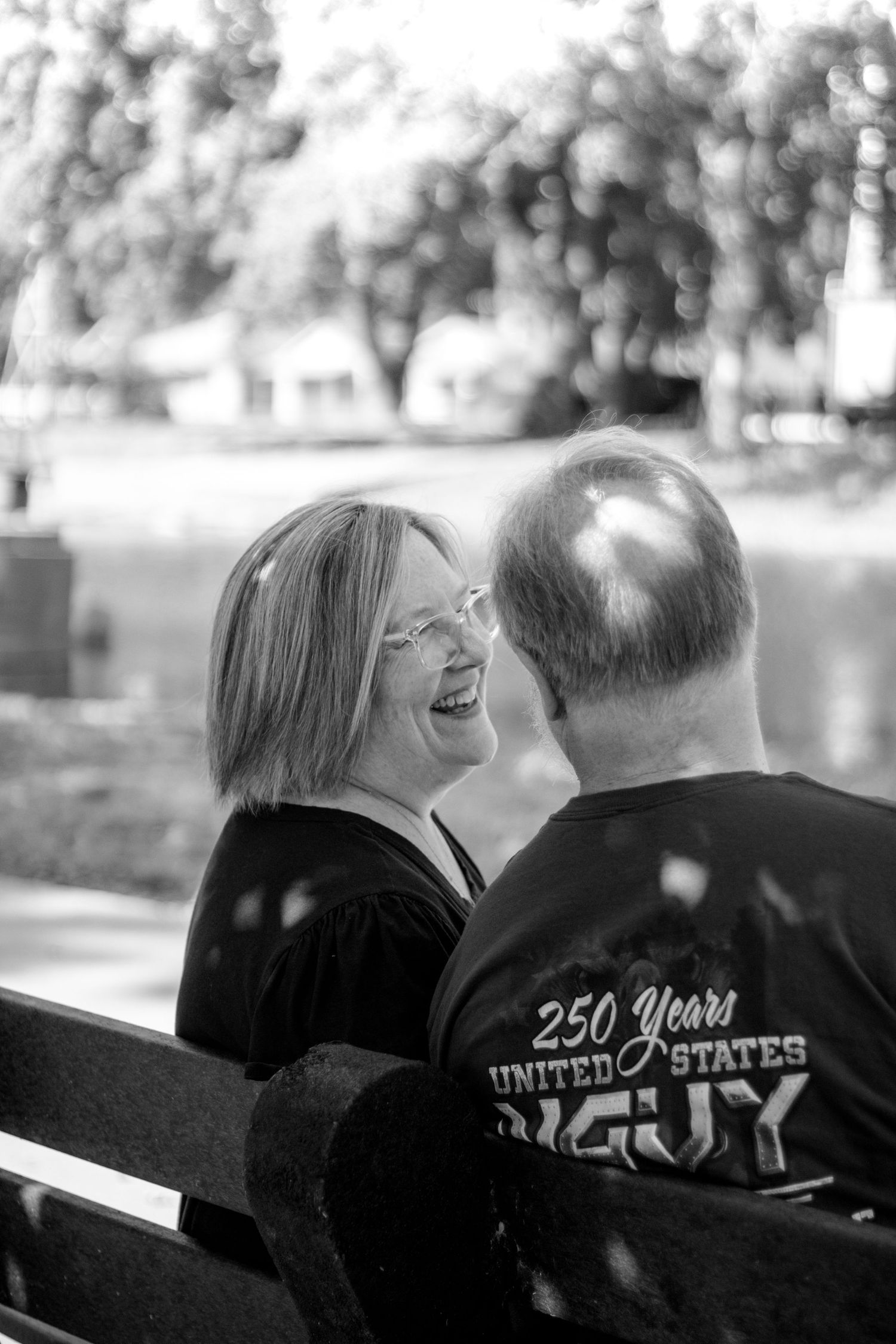 Black and white photo of a laughing woman and bald man sitting on a bench outdoors, wearing a 250 Years United States shirt.