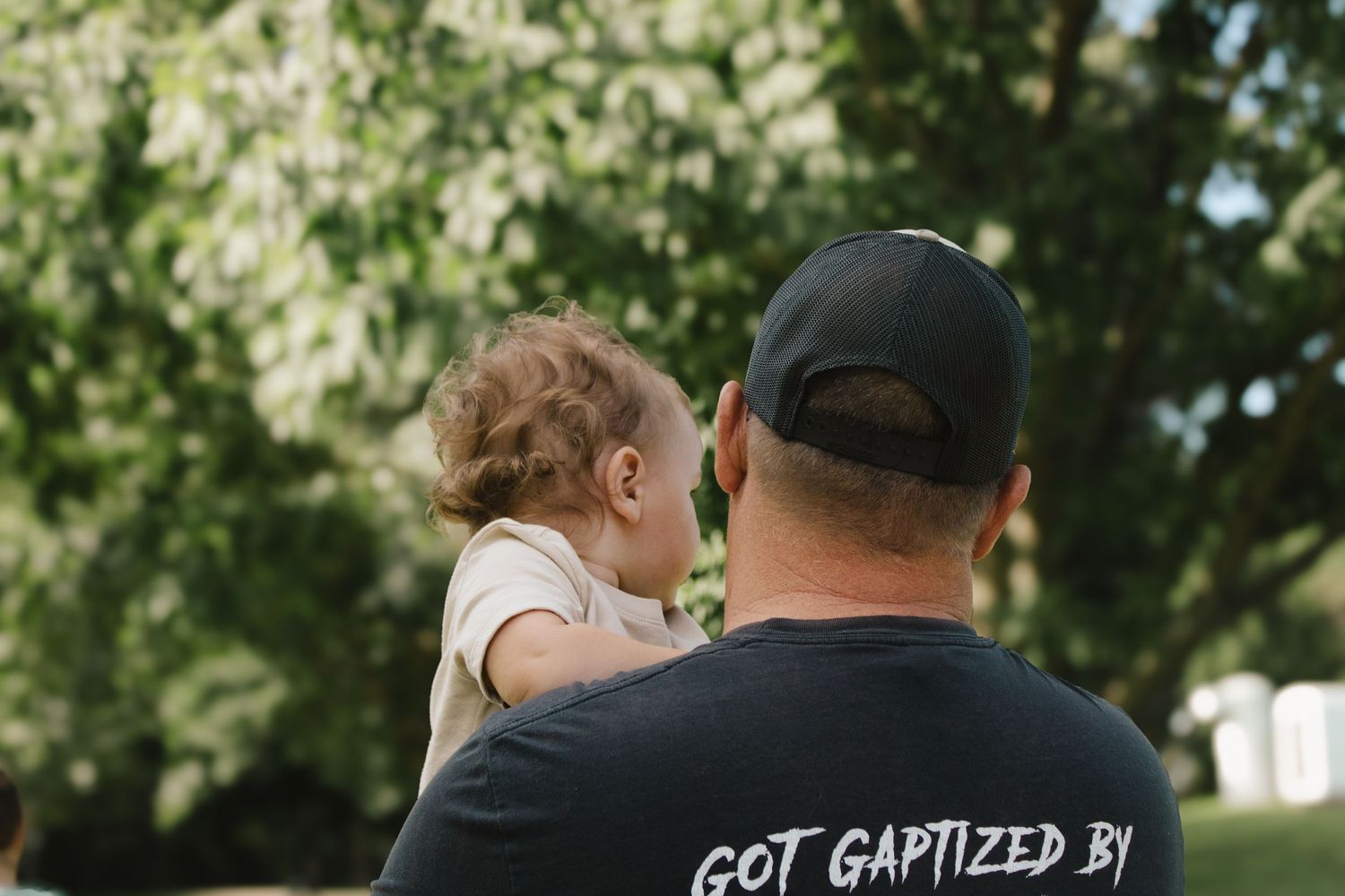 Father holding a toddler on his shoulder wearing a black Got Baptized t-shirt outdoors.