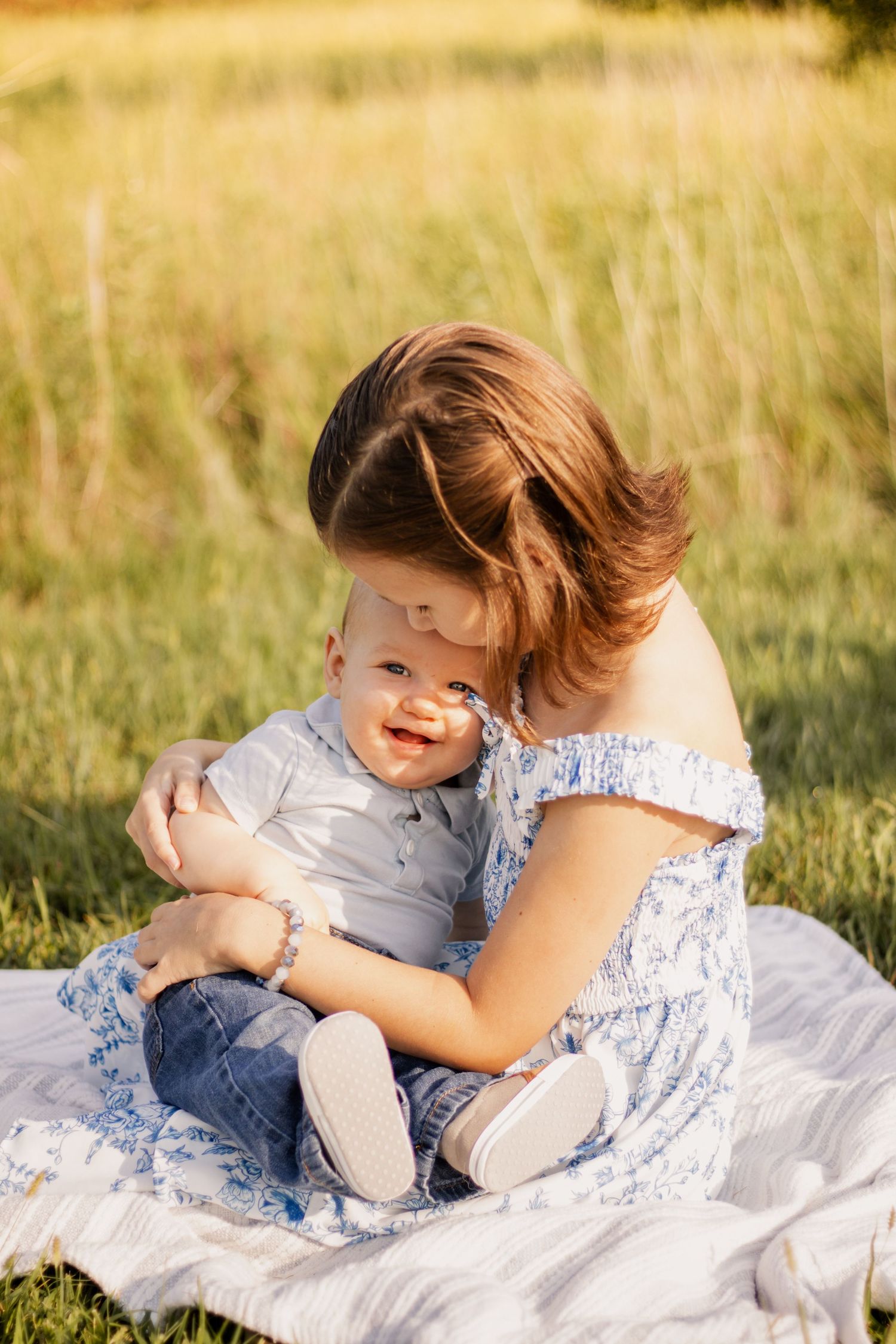 Mother in floral dress hugging smiling baby on blanket in sunny green field.