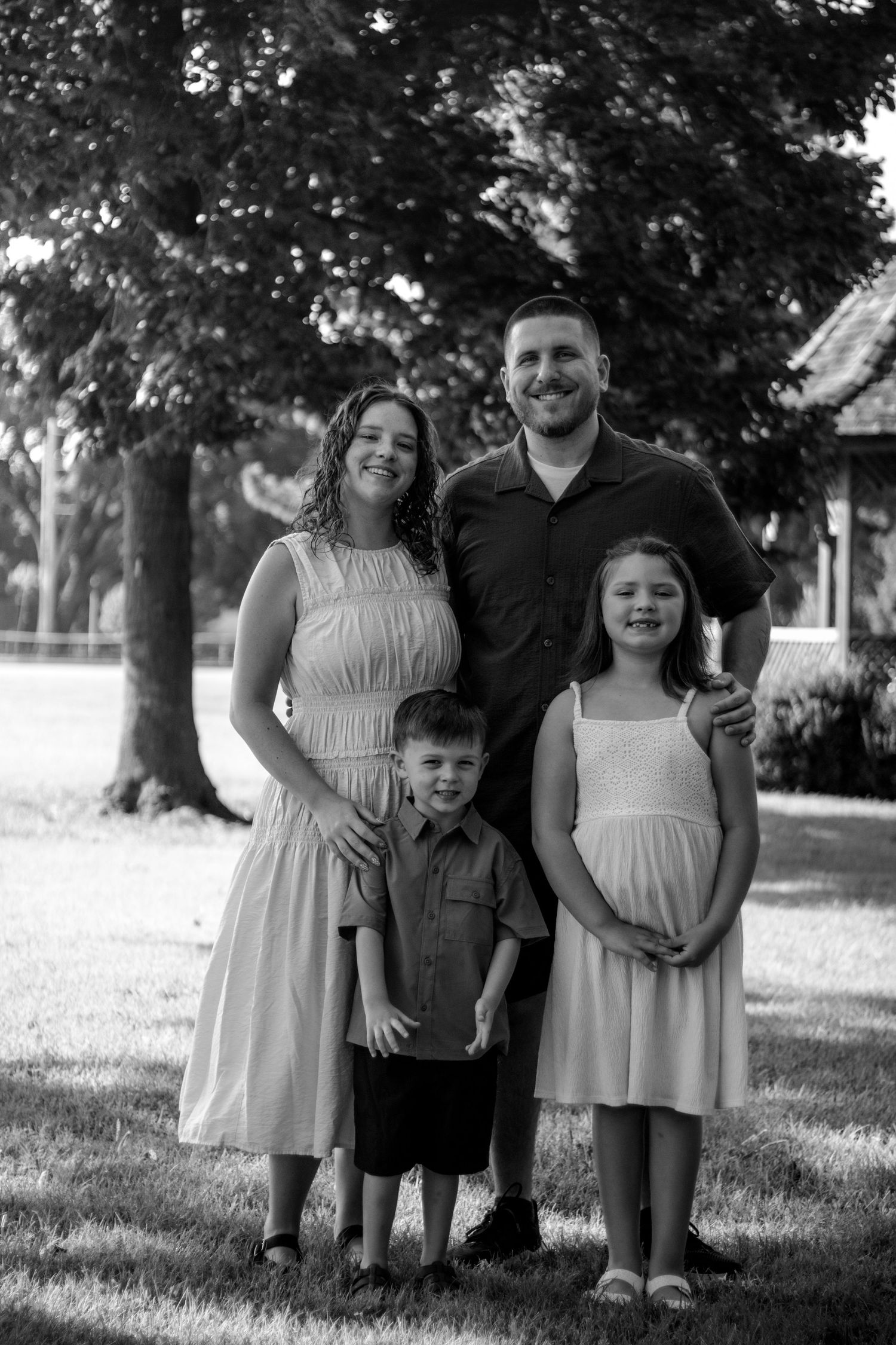 Black and white family portrait of parents with two children smiling outdoors under large trees.