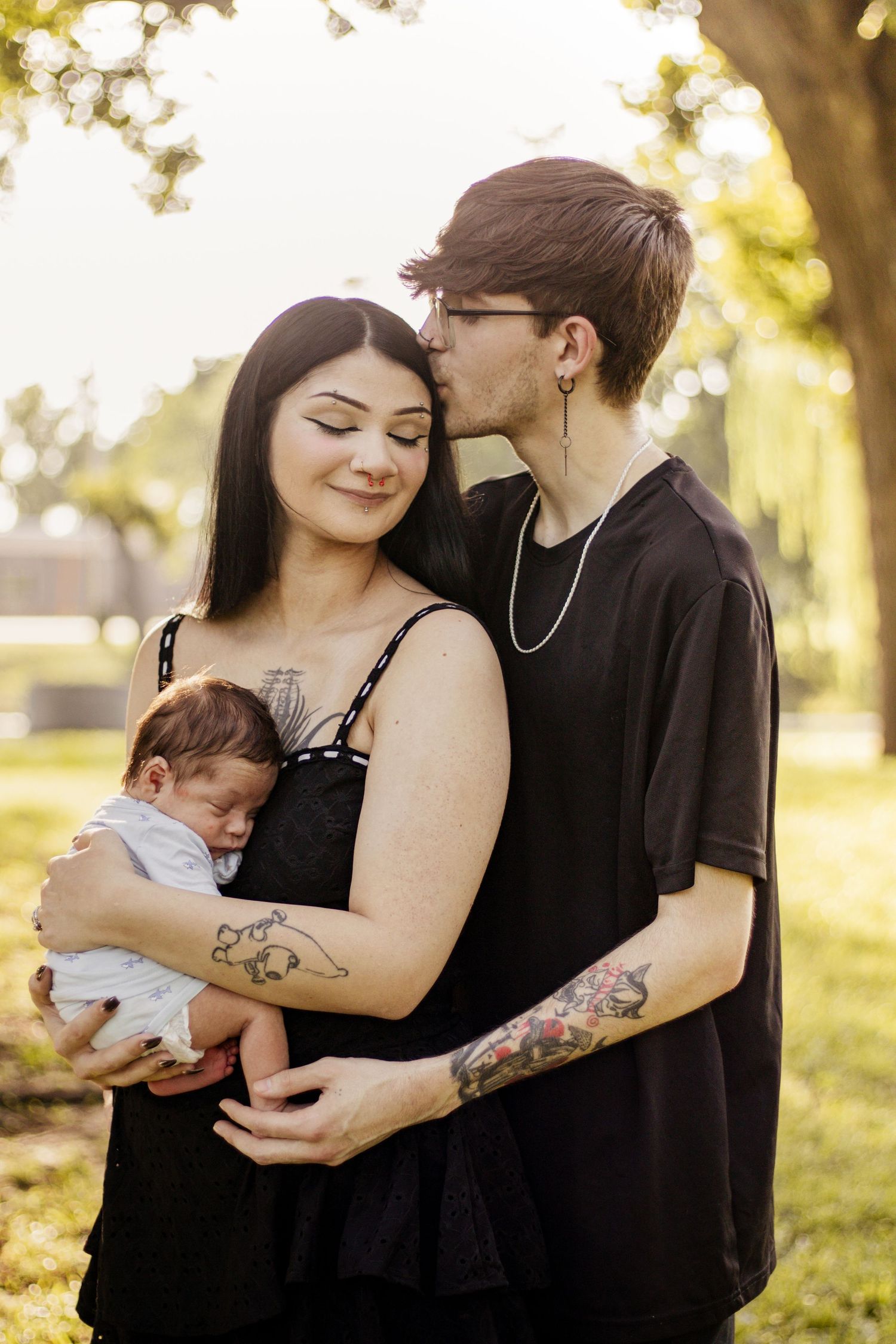 Young tattooed couple in black outfits holding newborn baby outdoors in sunny park setting.