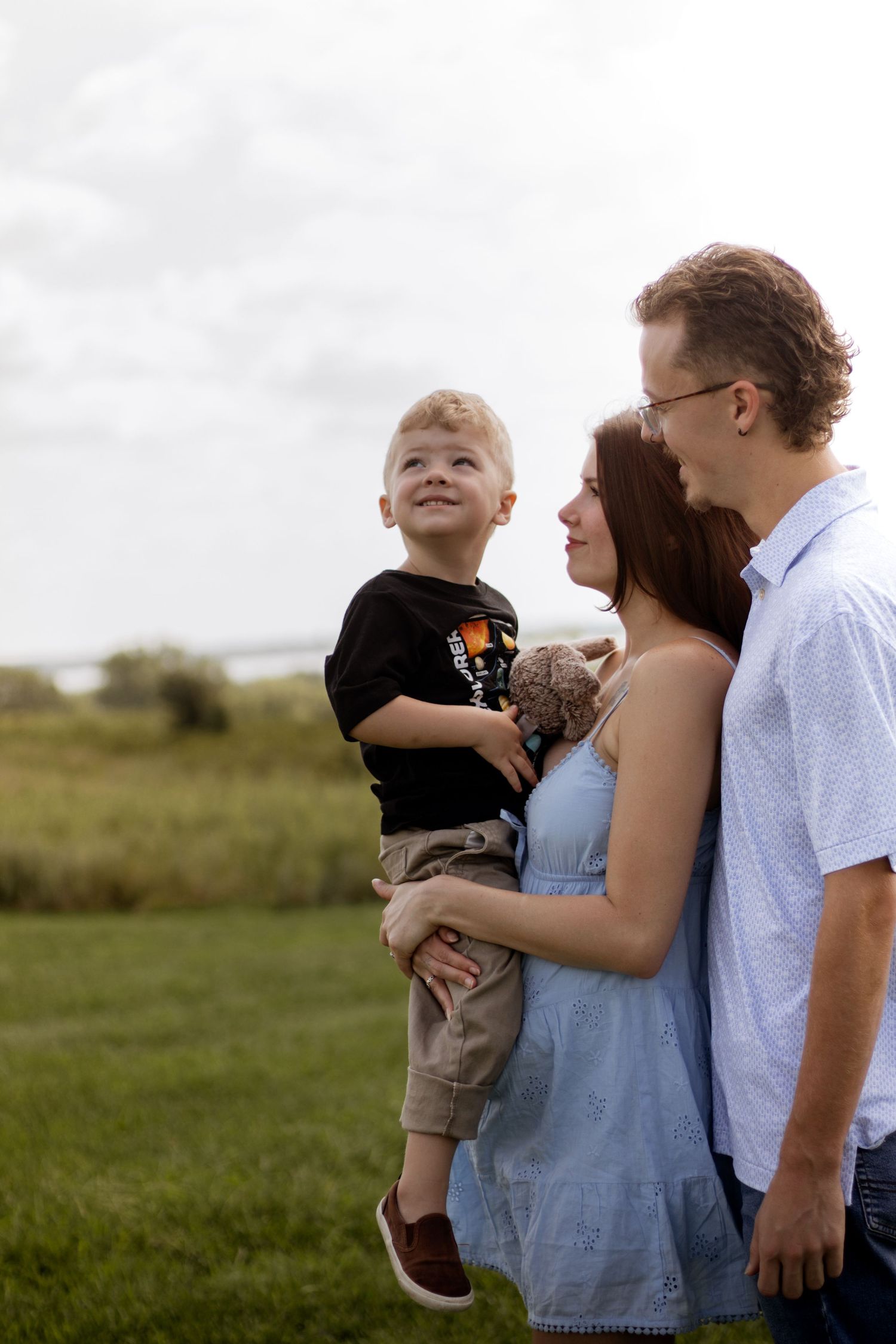 A young family of three stands outdoors, with parents holding their smiling toddler son carrying a stuffed bear.