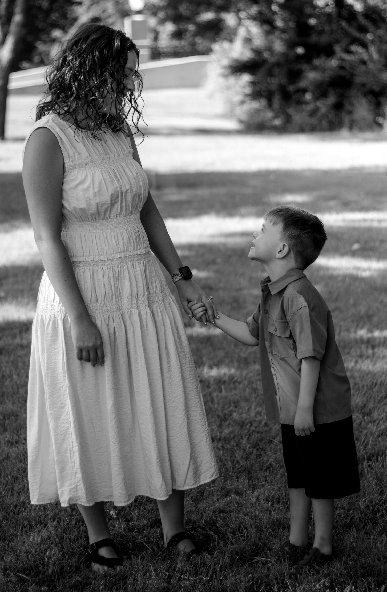 Black and white photo of a woman in a white dress holding hands with a young boy in a park setting.