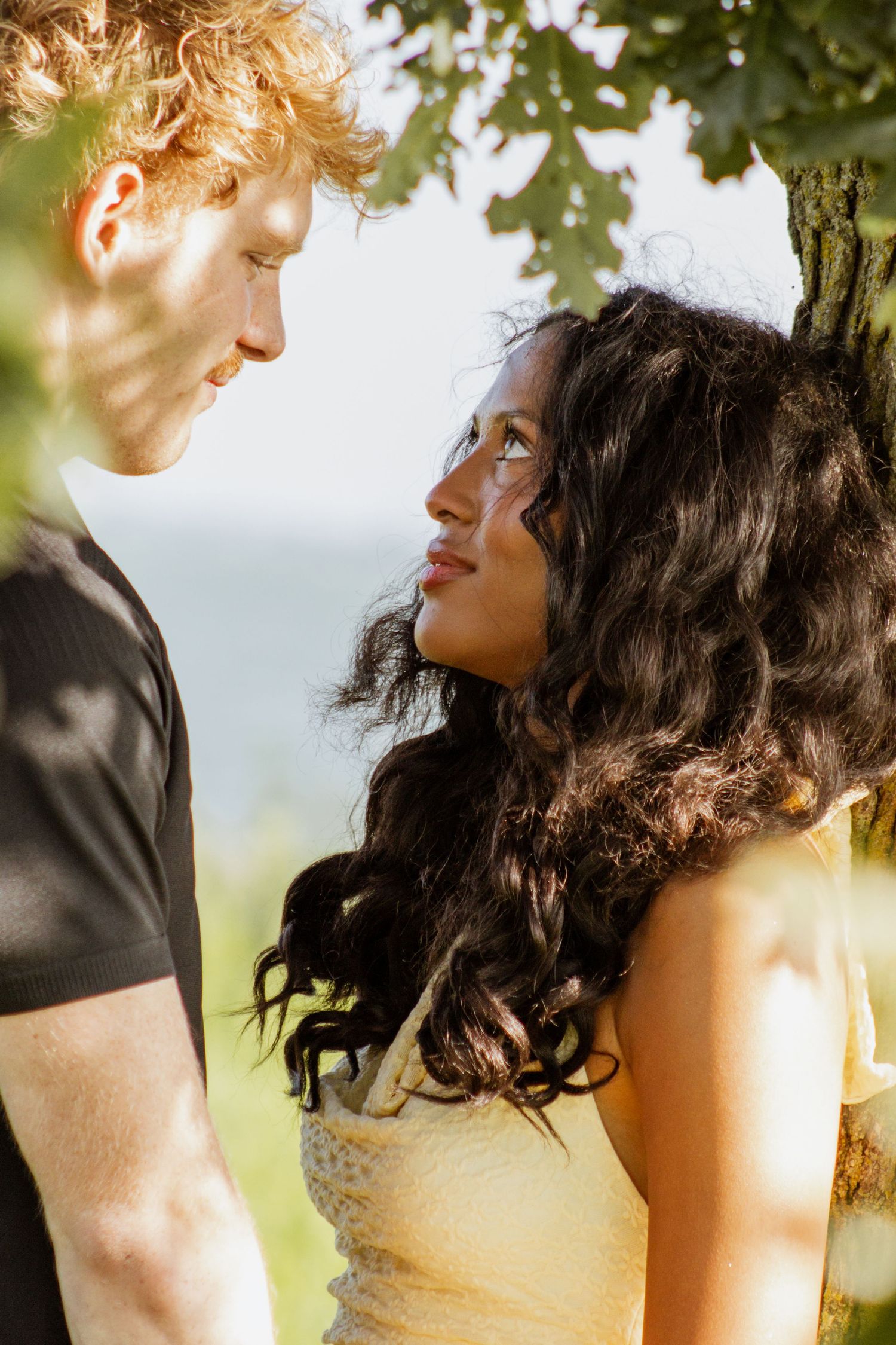 Romantic interracial couple gazing at each other under a tree, man with curly red hair, woman with long dark hair.