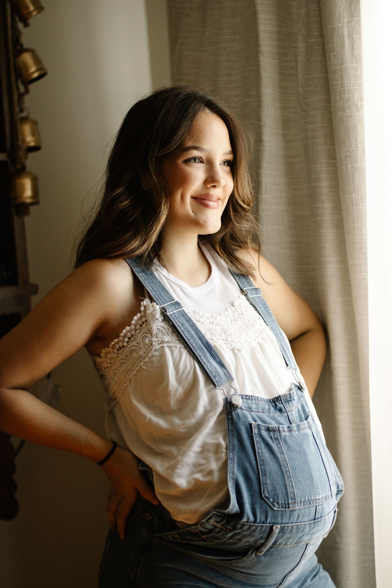 Smiling pregnant woman in denim overalls and white lace top standing by a window in natural light.