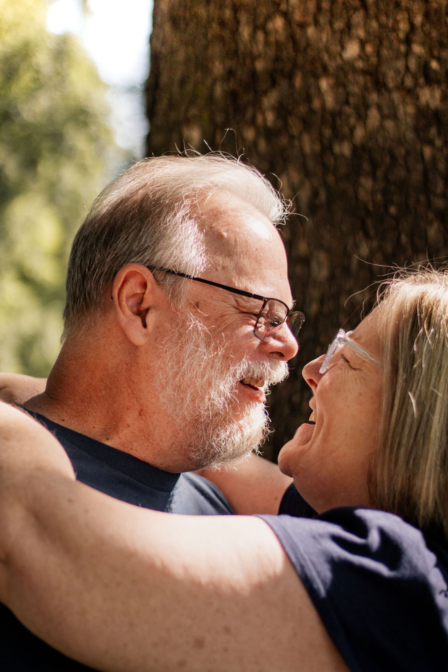 An elderly couple embracing and laughing together outdoors near a tree on a sunny day.