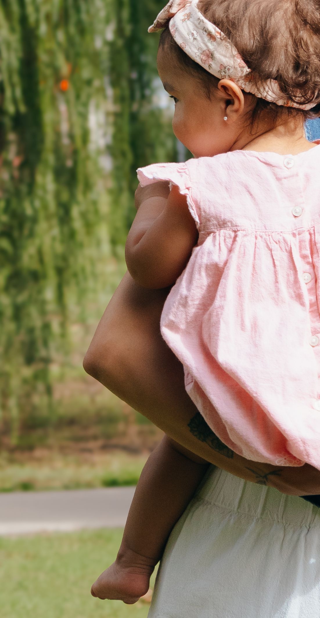 A woman holds a baby girl in a pink dress and floral headband outdoors near lush green willow trees.
