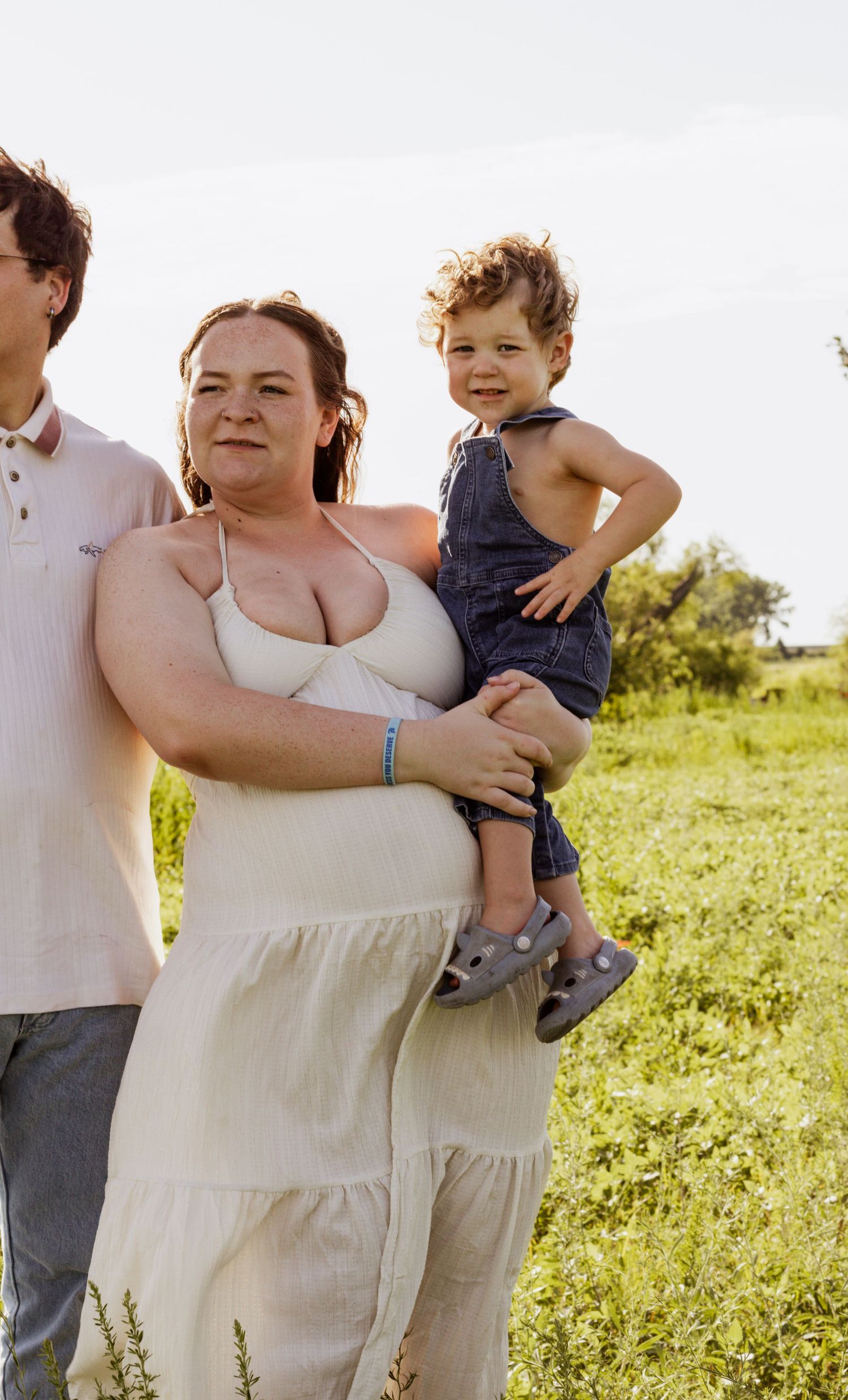 Pregnant woman in white dress holds toddler in denim overalls outdoors in a sunny green field with family.
