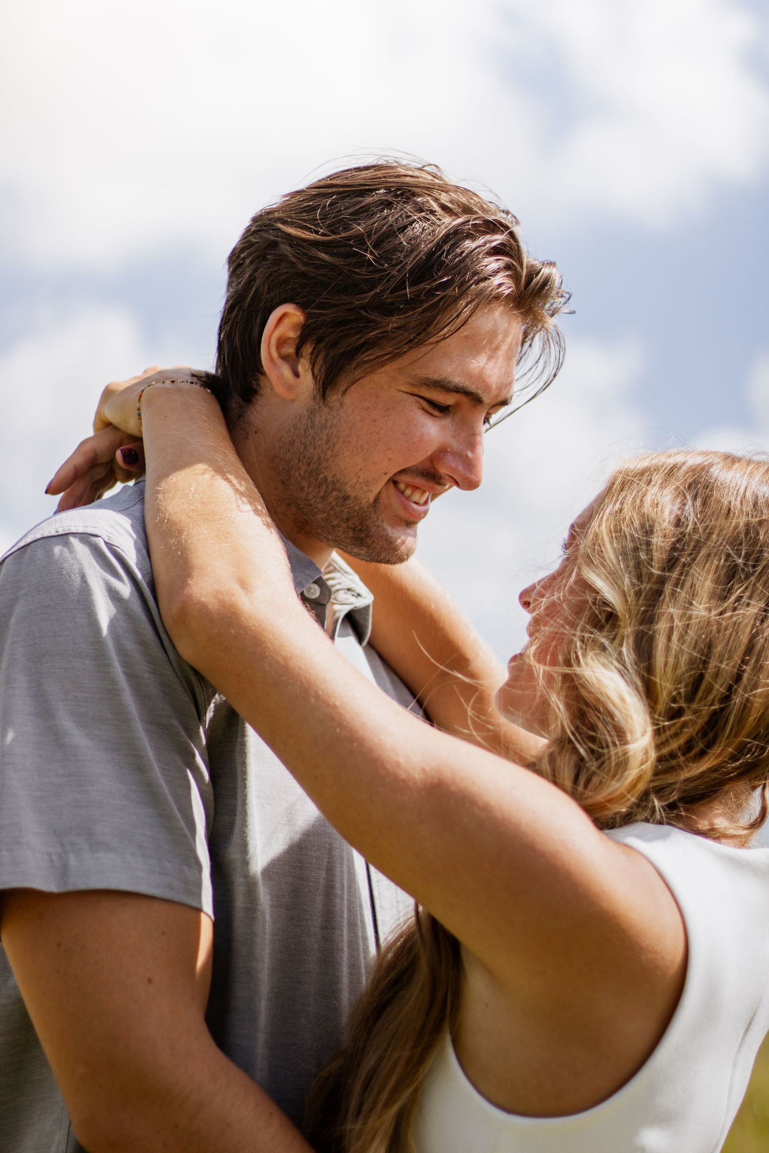 Smiling couple embracing outdoors under a cloudy sky, sharing a romantic moment face to face.