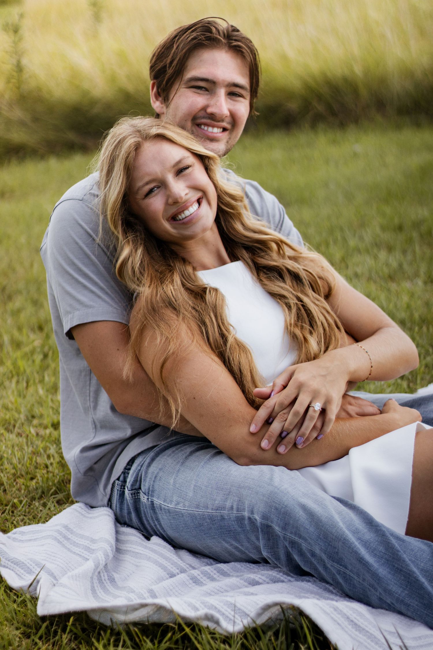 Happy couple sitting on blanket in grass, woman with long blonde hair embraced by smiling man in gray shirt.
