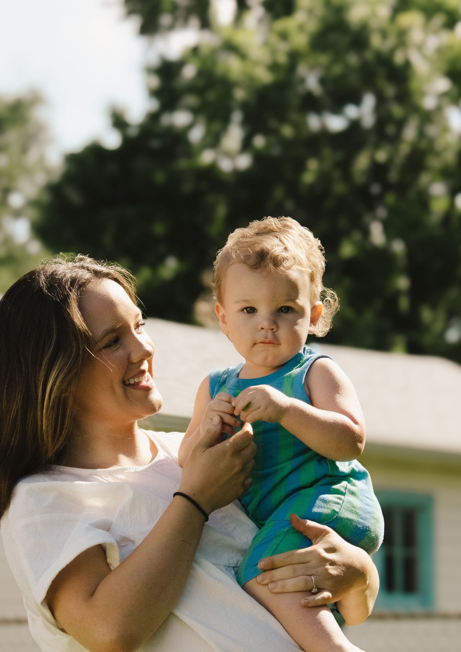 Smiling mother in white top holding her toddler in blue outfit outdoors.