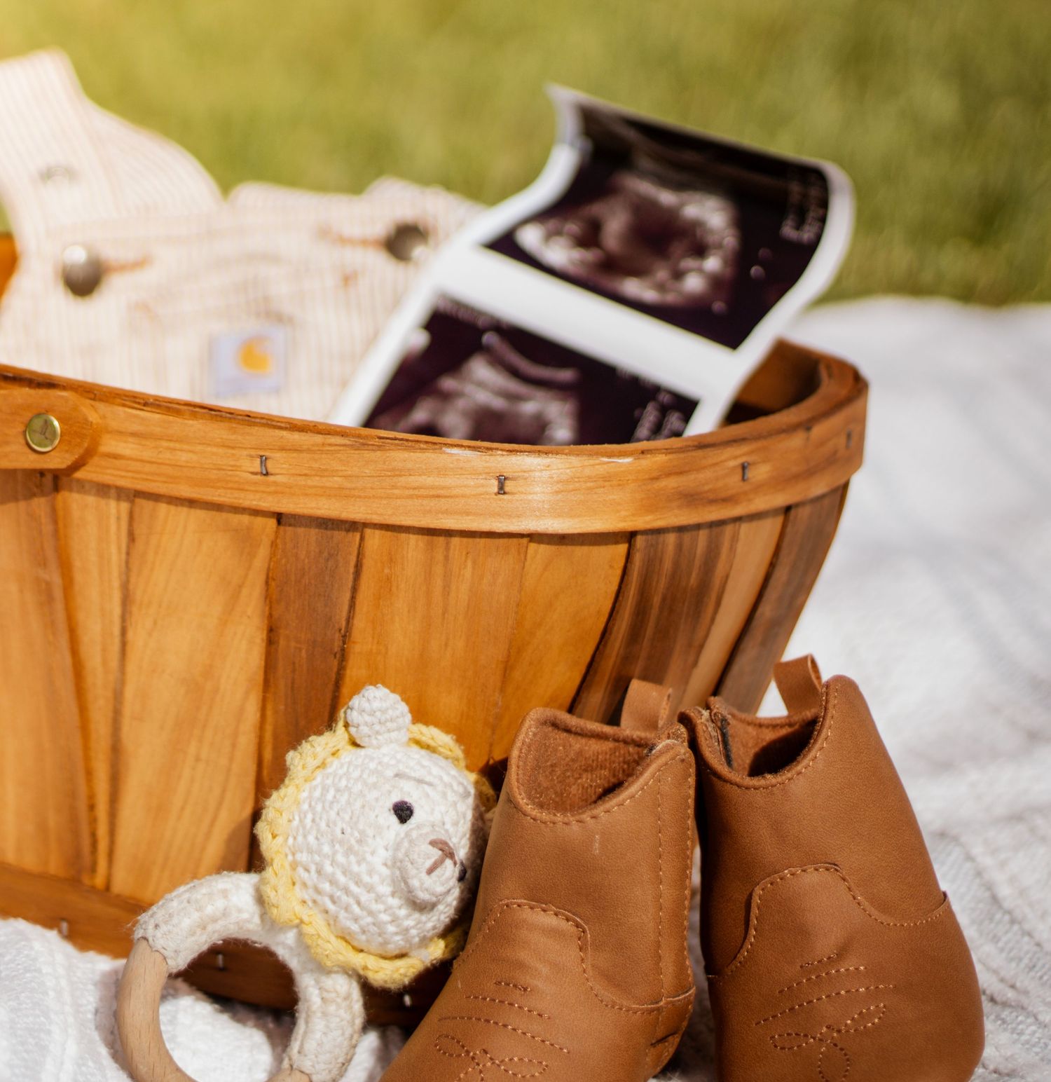 Wooden basket with baby ultrasound photos, brown leather booties, and a crocheted bear toy on a white blanket outdoors.