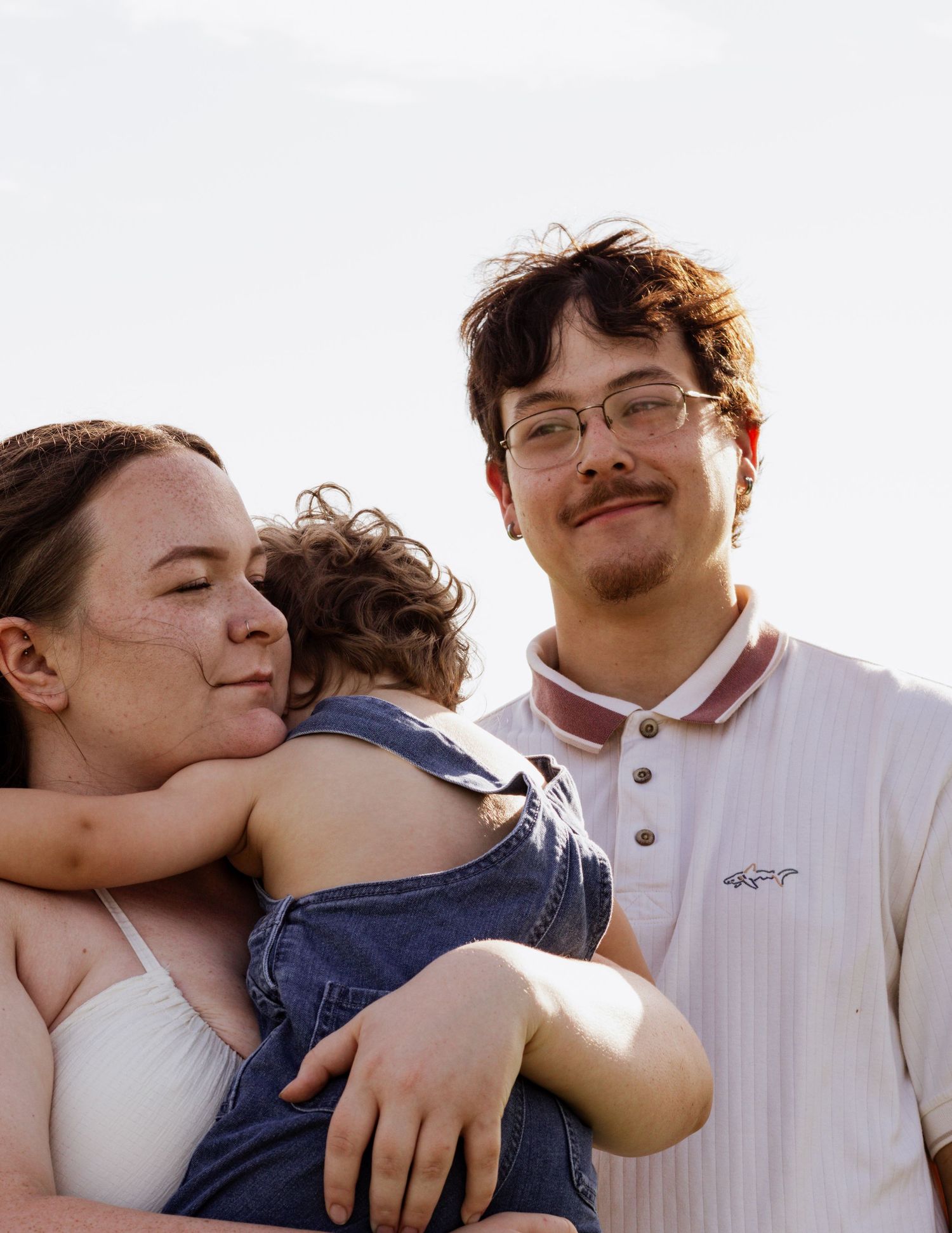 A young couple smiling and holding a toddler in denim overalls outdoors against a bright sky.