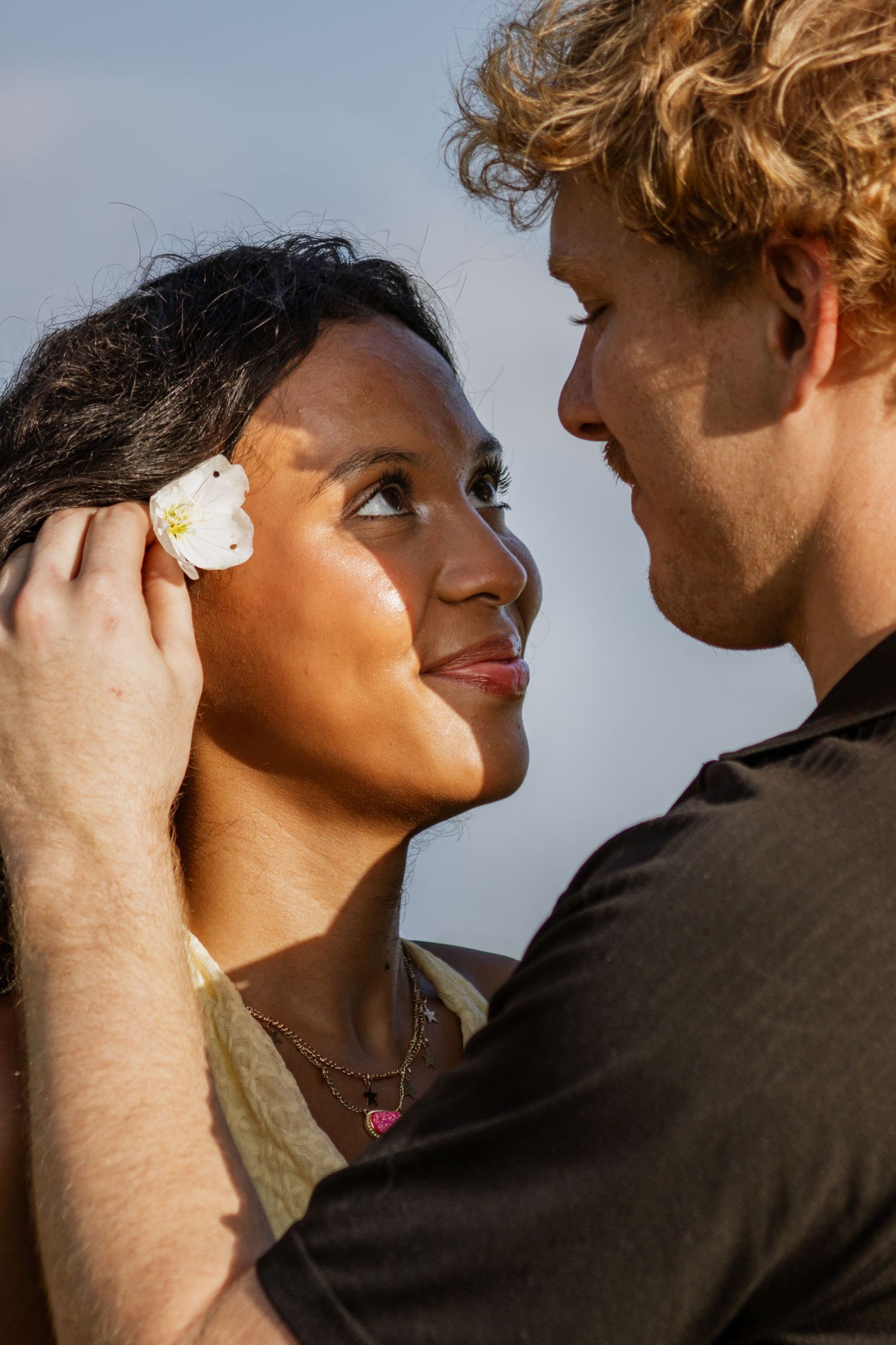 Romantic interracial couple gazing at each other, man tucking a white flower behind womans ear outside.