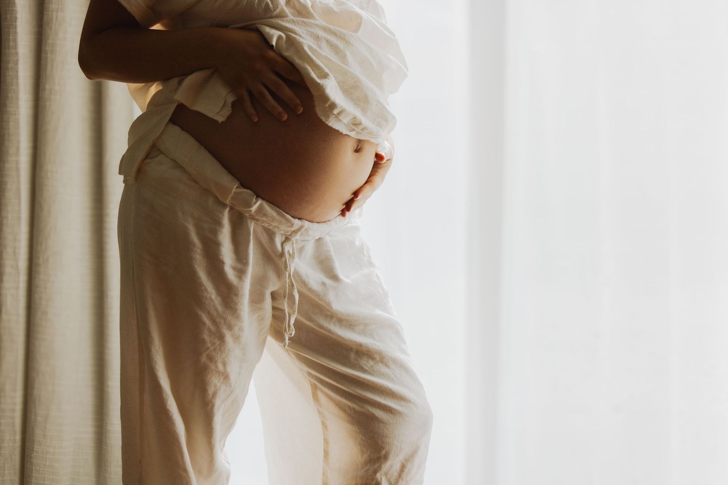 Pregnant woman in white linen top and pants holds baby bump beside softly lit curtained window.