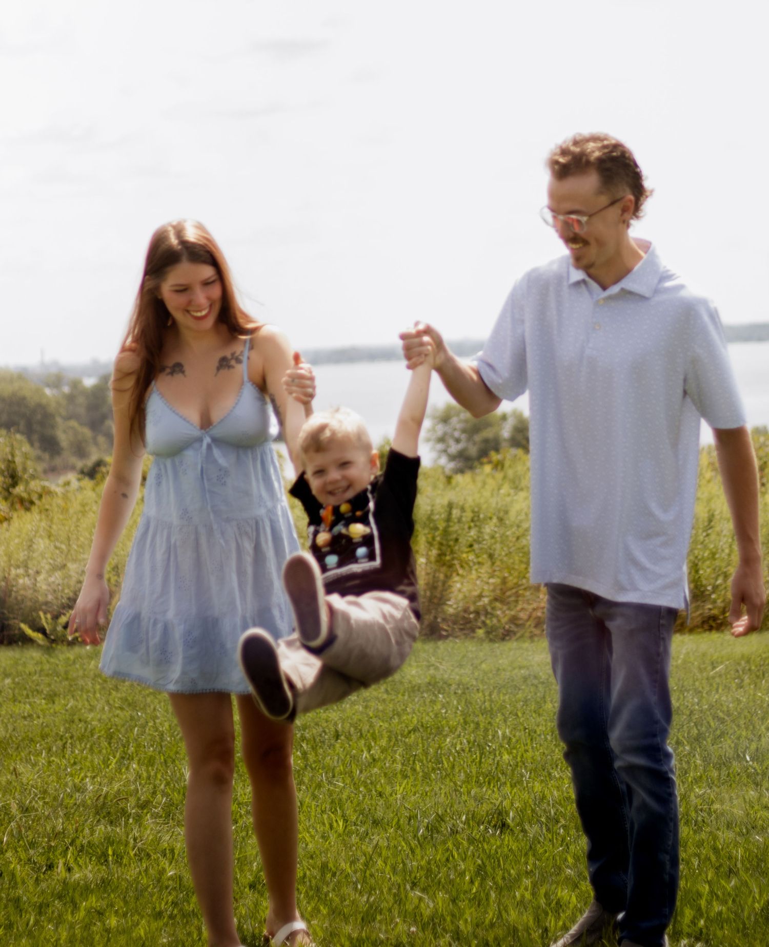 A happy family swings their young child between them while walking on green grass near a lake.
