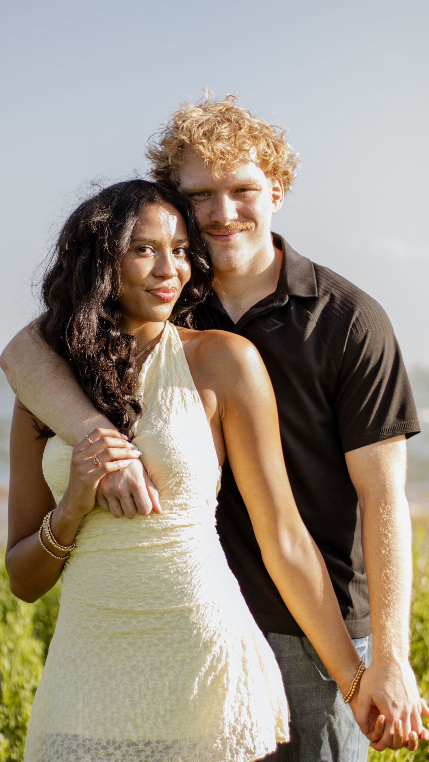 Happy interracial couple embracing outdoors, woman in white halter dress with gold bracelets, man in black polo shirt.
