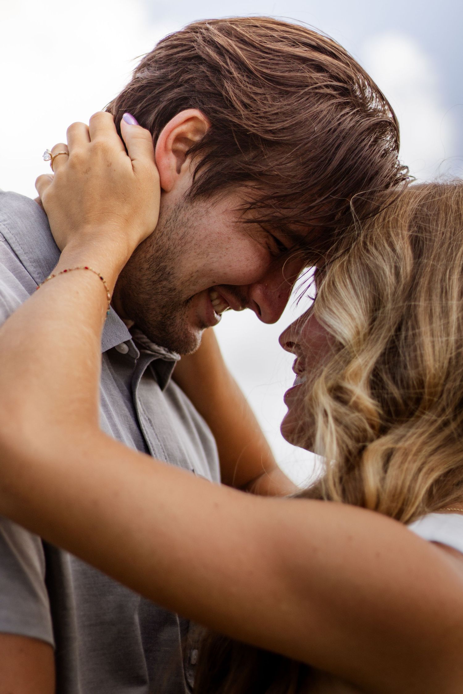 Romantic couple smiling and embracing outdoors, woman holding man's head tenderly under a cloudy sky.
