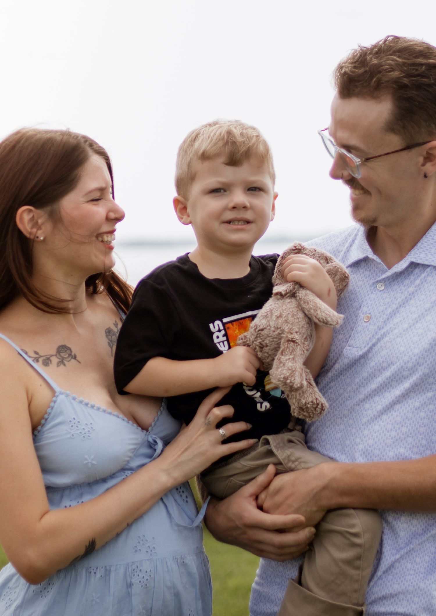 A happy family of three, with parents holding their young blonde boy clutching a stuffed teddy bear outdoors.