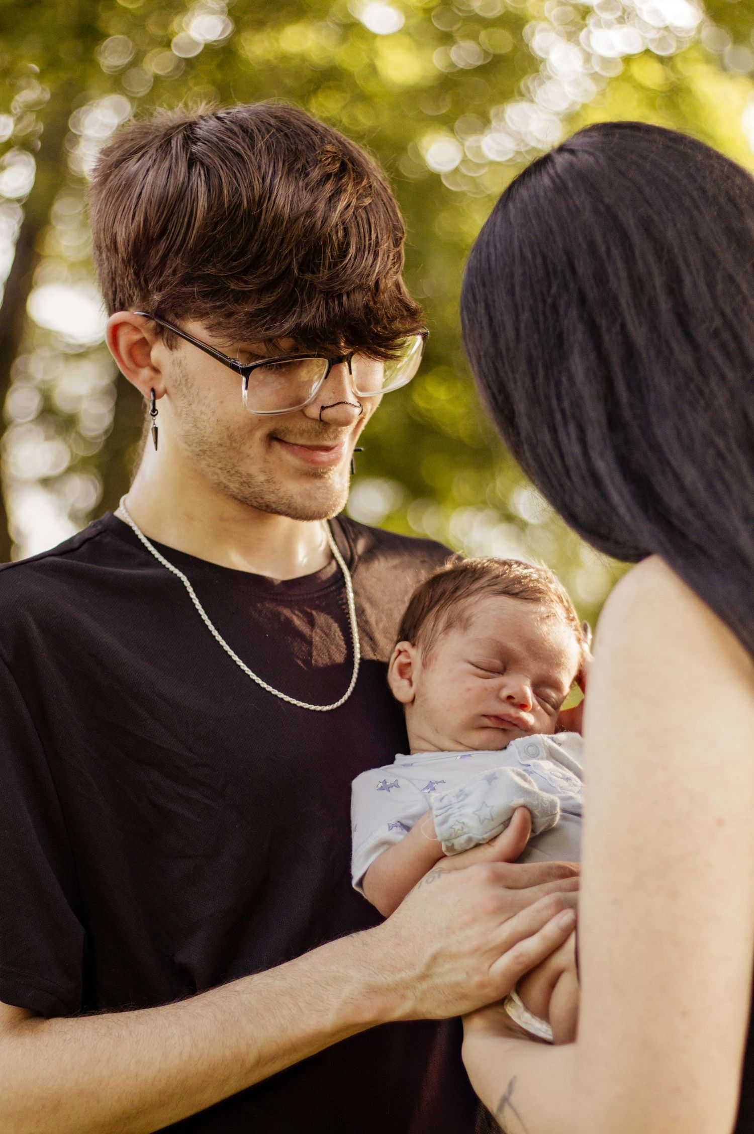 A young man wearing glasses and a black t-shirt smiles while holding a sleeping newborn baby outdoors.