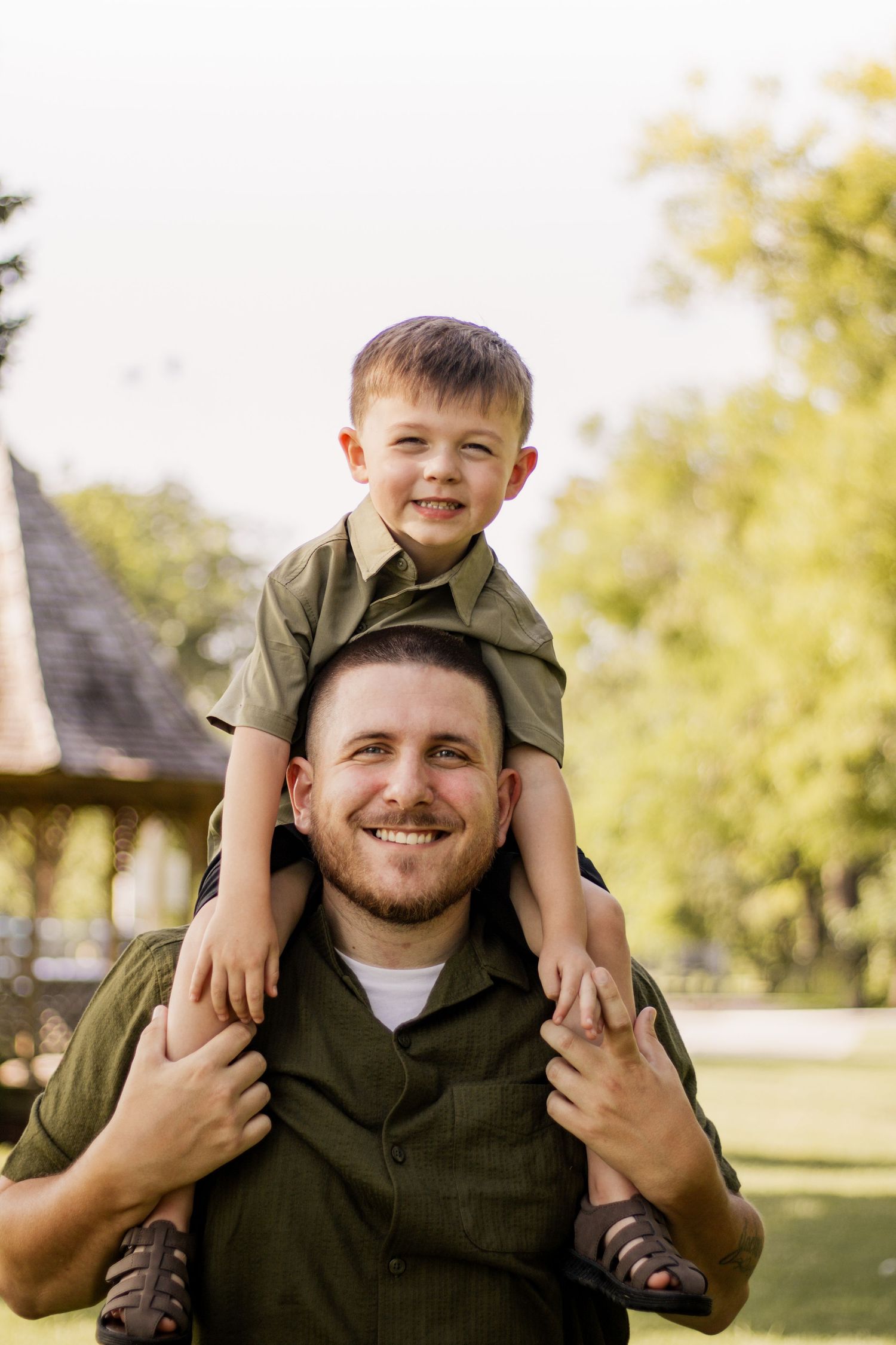 A smiling father carries his young son on his shoulders in a sunny green park, both wearing matching olive green shirts.