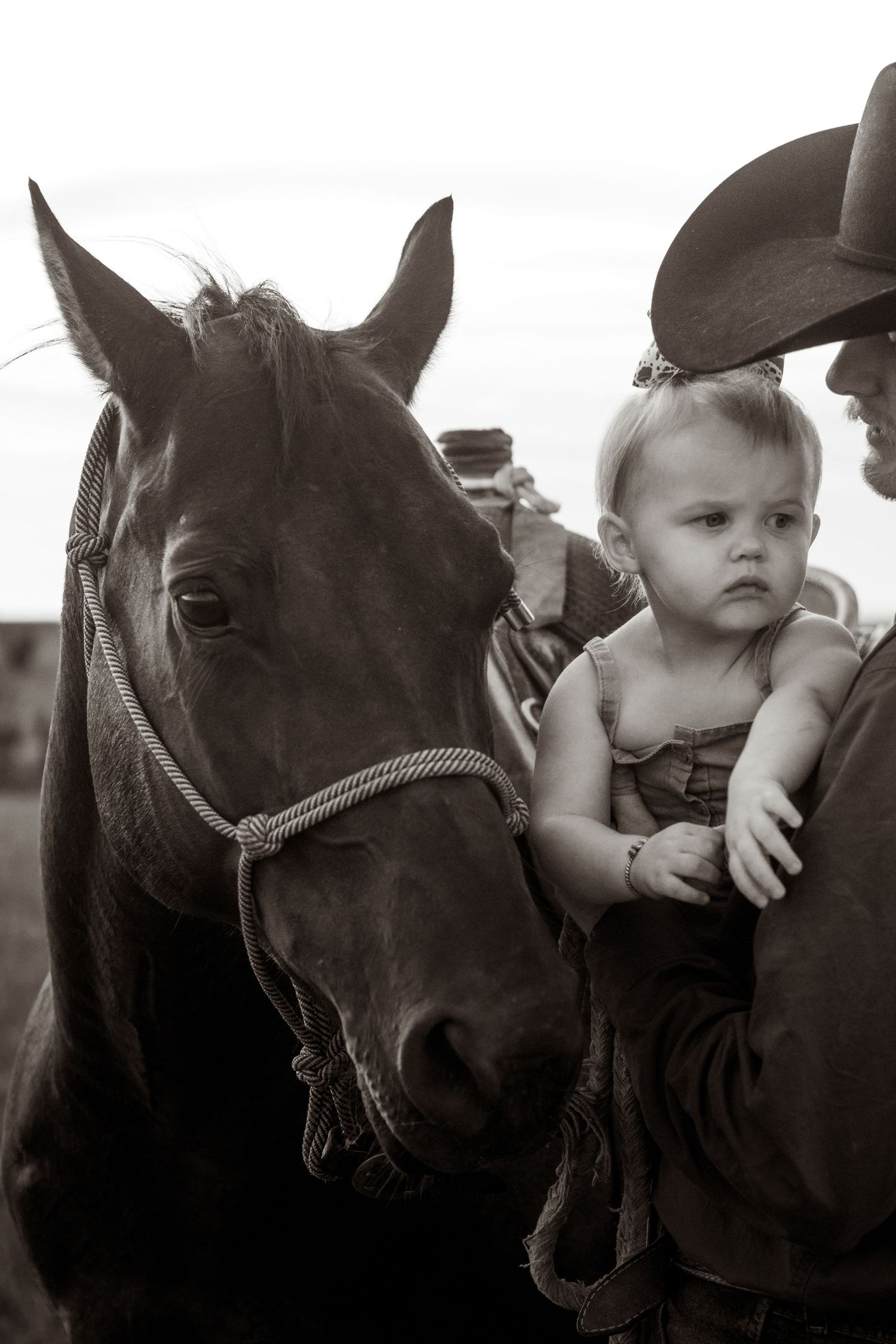Black and white photo of a baby girl being held next to a dark horse by a cowboy.