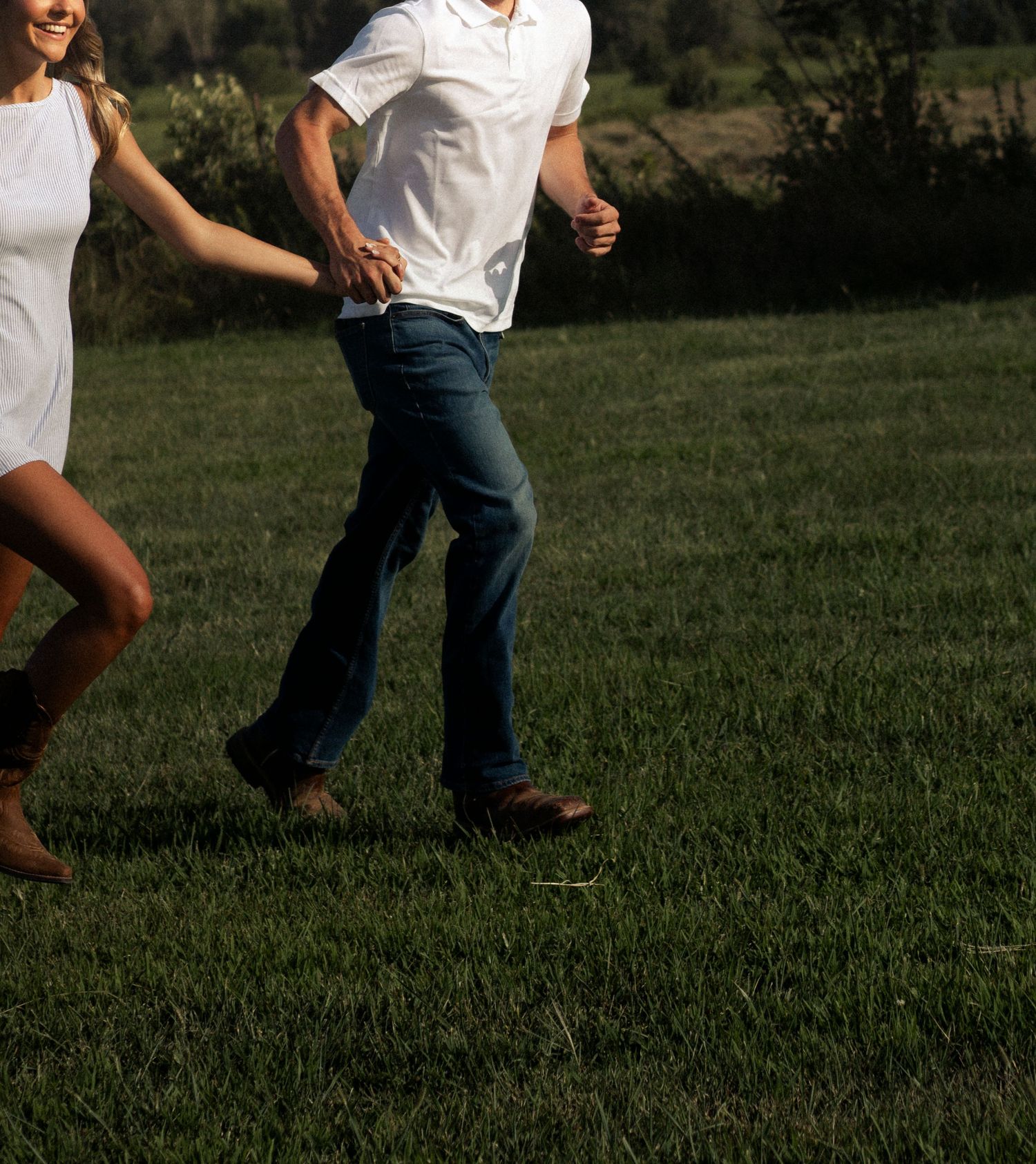 Couple holding hands running through a lush green field, wearing white shirts and blue jeans on a sunny day.