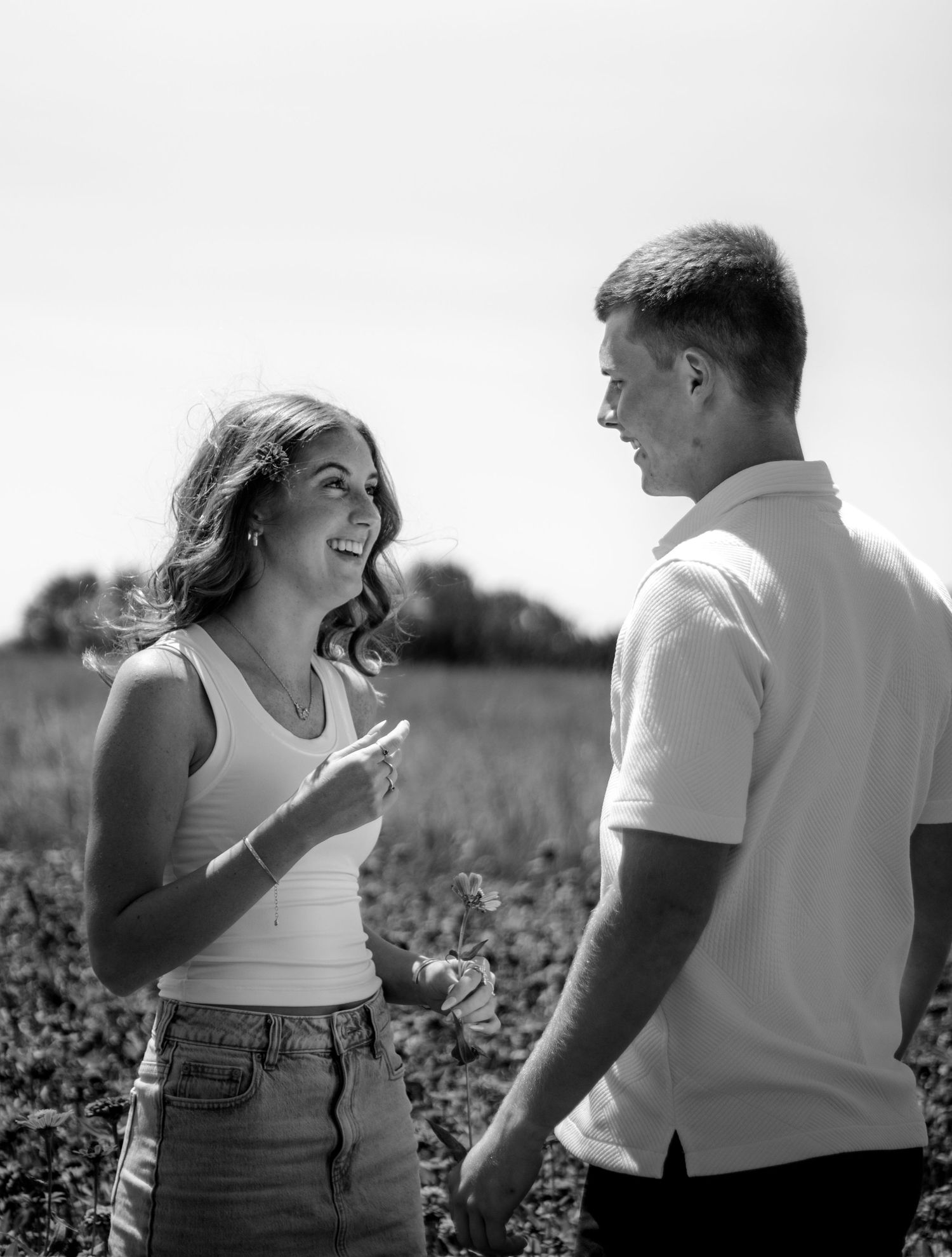 Black and white photo of a smiling couple talking in a sunny field, woman in tank top and denim skirt.