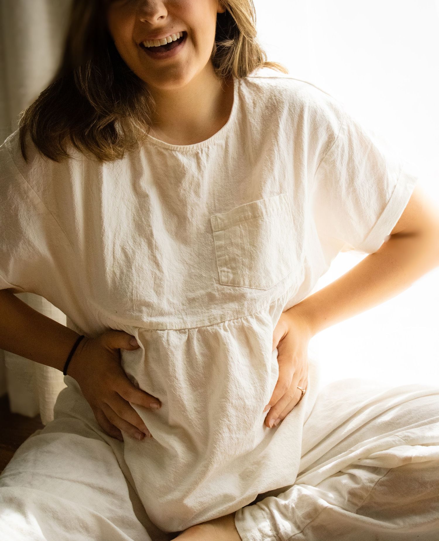 Smiling pregnant woman in white linen top sitting on bed cradling her baby bump with both hands.