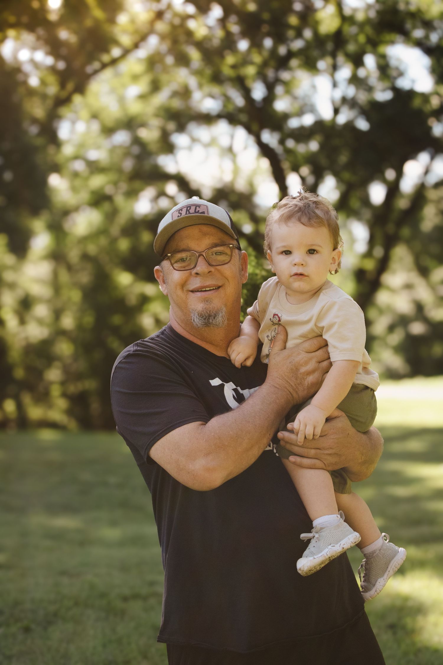 Middle-aged man in SRC black t-shirt and cap holding a young toddler in a sunny park setting.